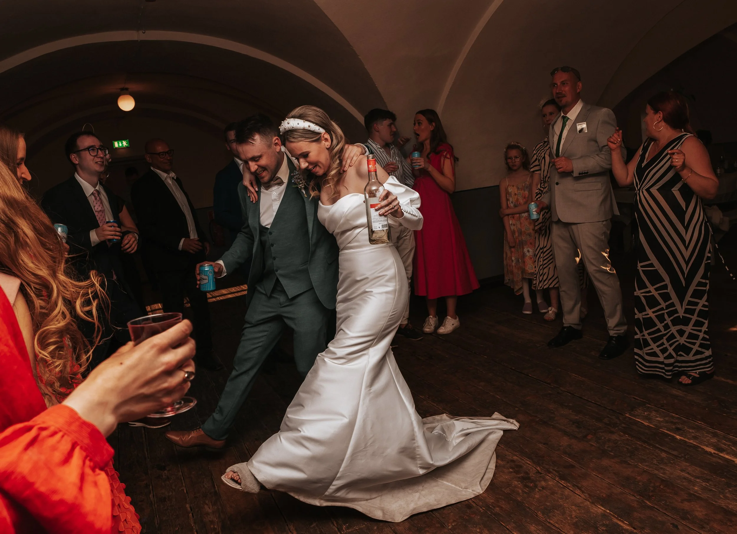 Wedding celebration with bride and groom dancing, surrounded by guests, in a dimly lit venue.