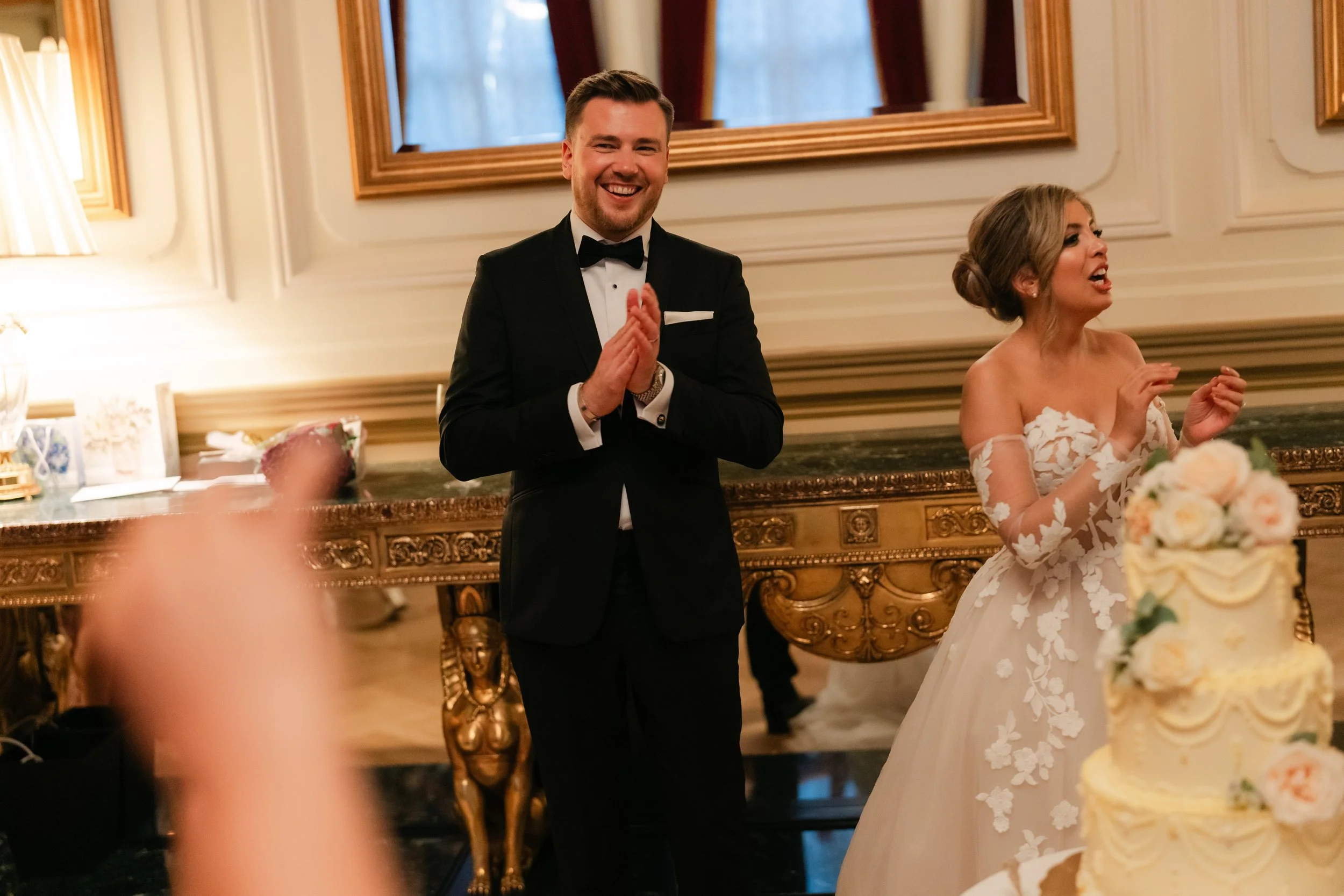 Bride and groom clapping and celebrating at a wedding reception, with a decorated cake in the foreground.