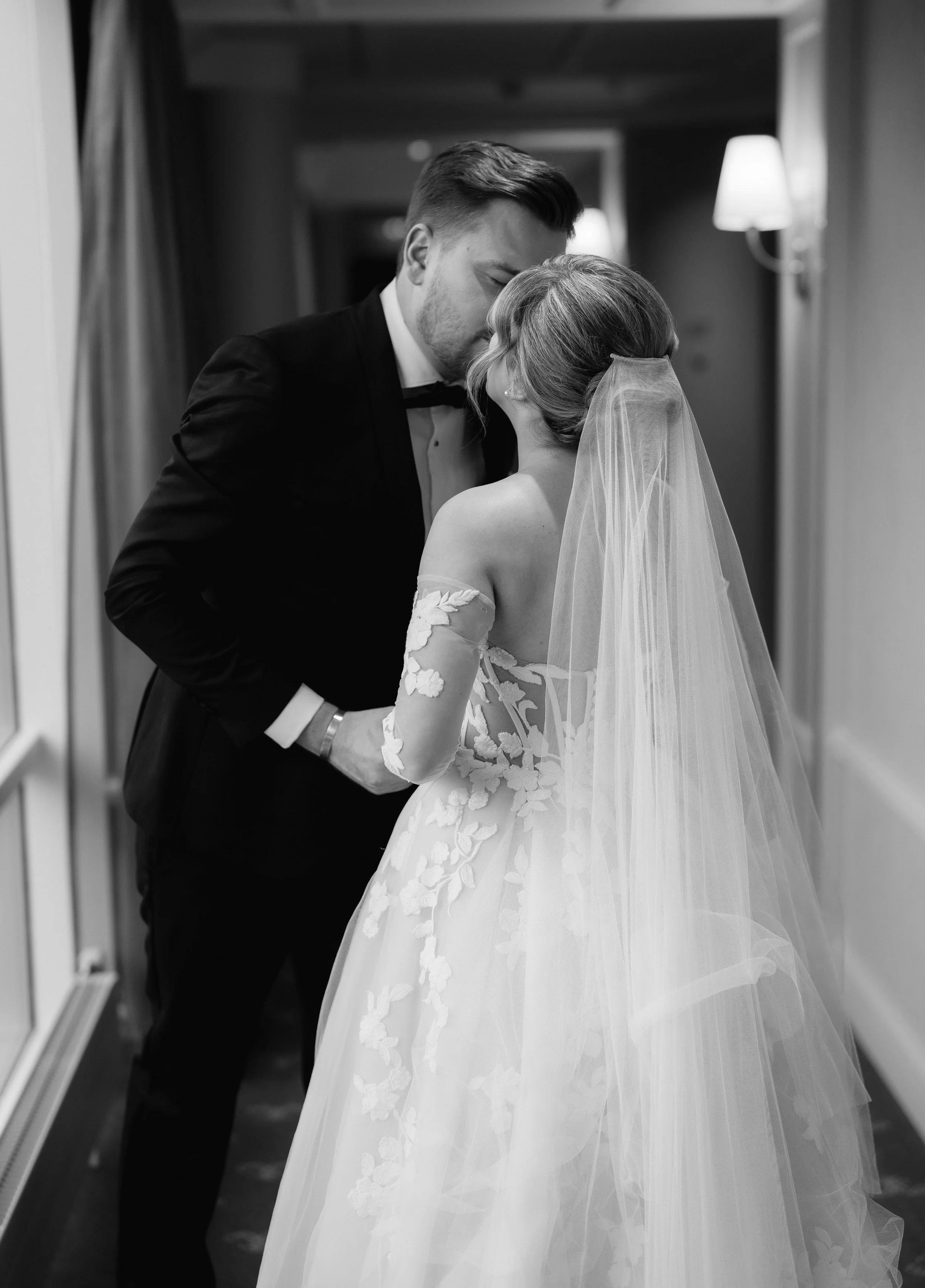 Bride and groom kissing in hallway, wedding attire, black and white photo.