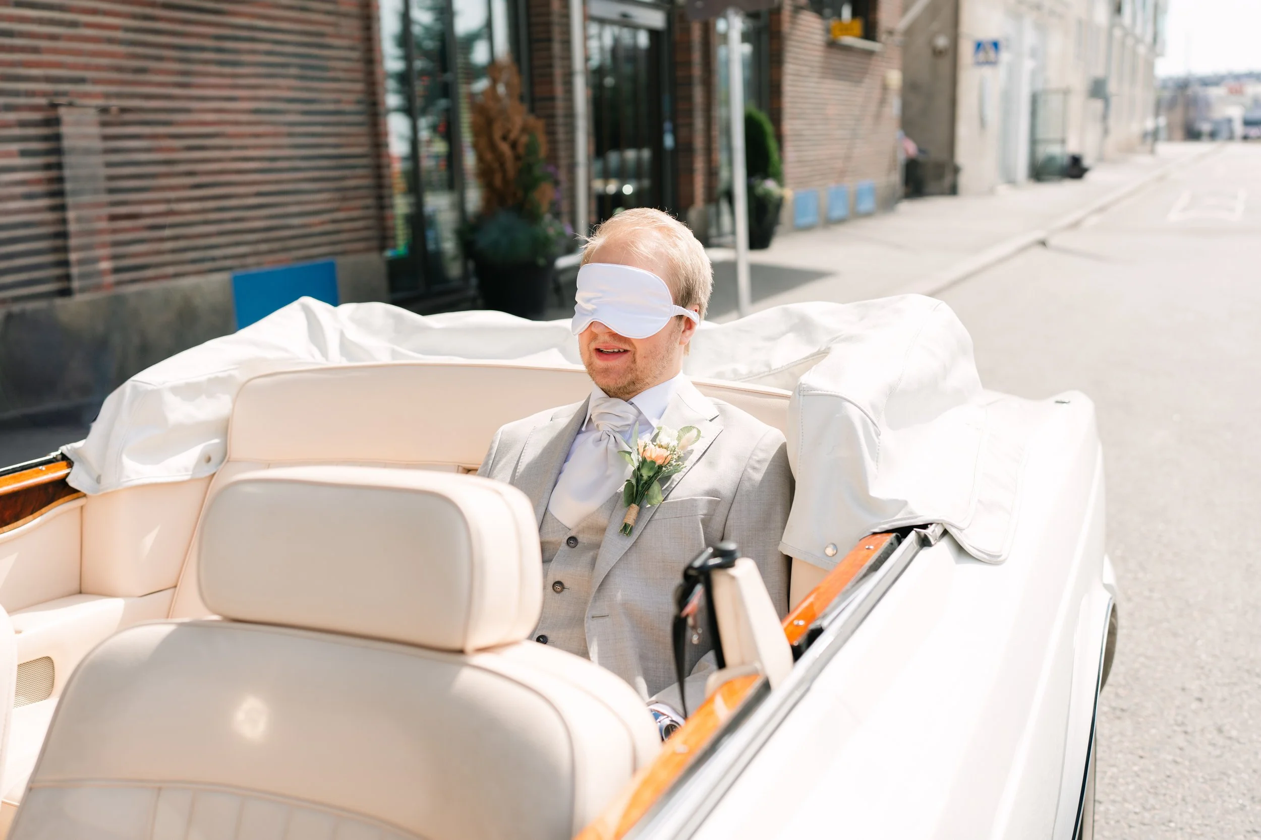 Blindfolded groom in convertible car wearing wedding attire with boutonniere.