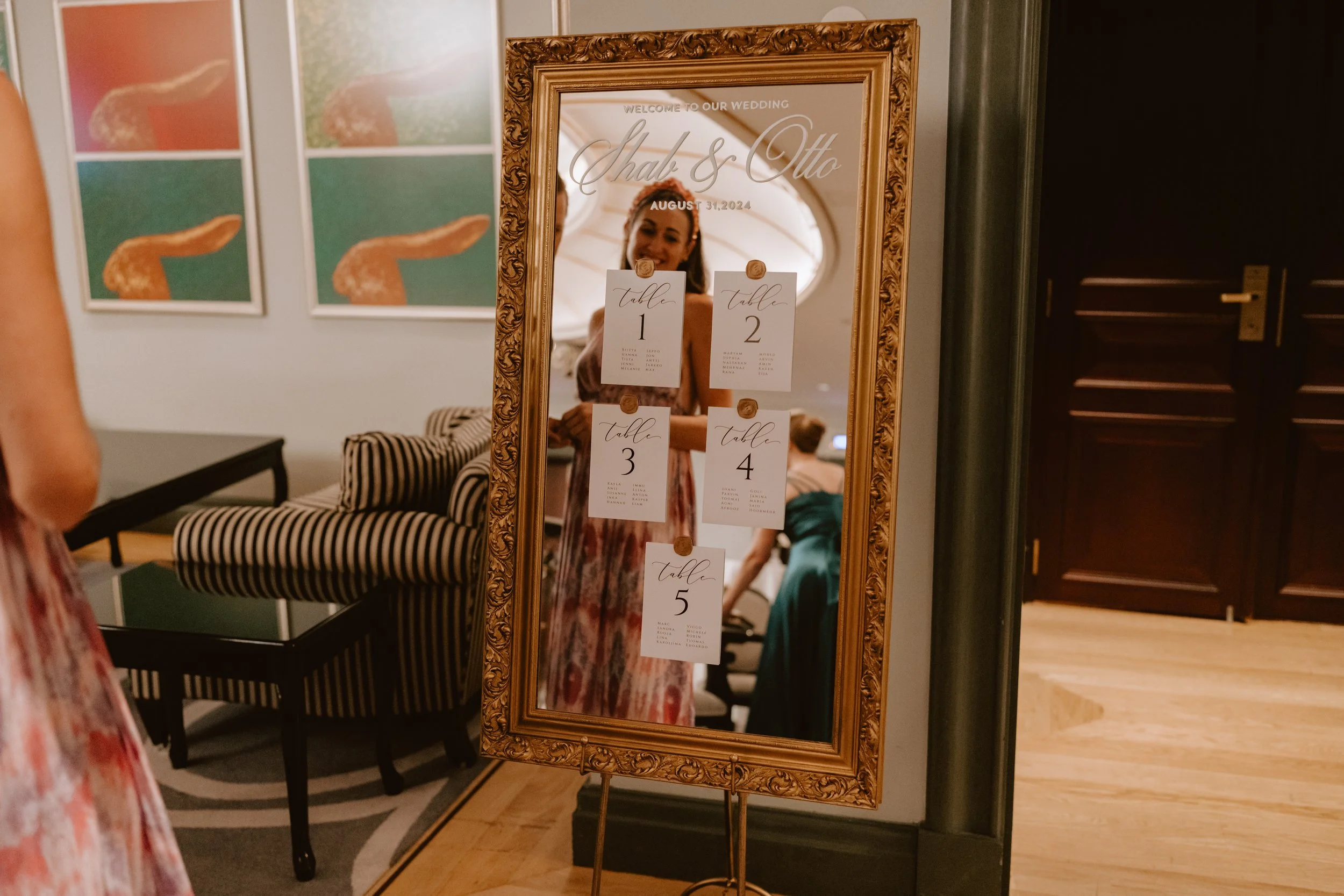 Elegant mirror with wedding seating chart in ornate frame, reflecting a woman in floral dress, indoor setting with modern decor.