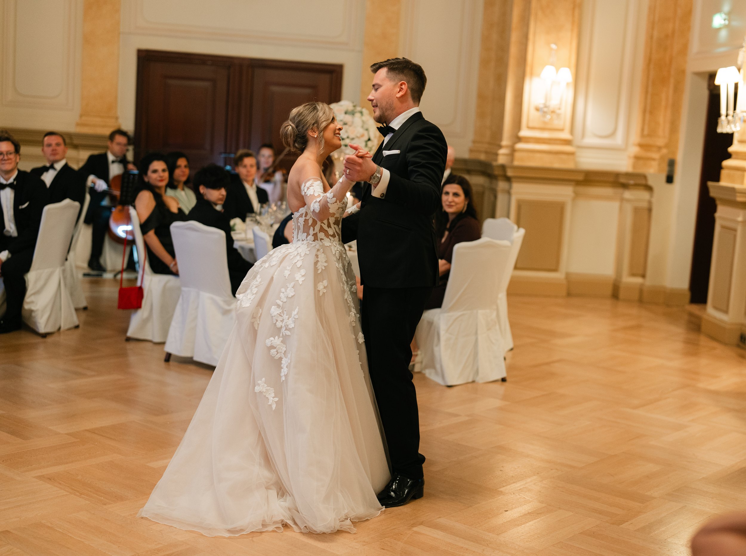Bride and groom dancing at wedding reception with guests seated, elegant ballroom setting.