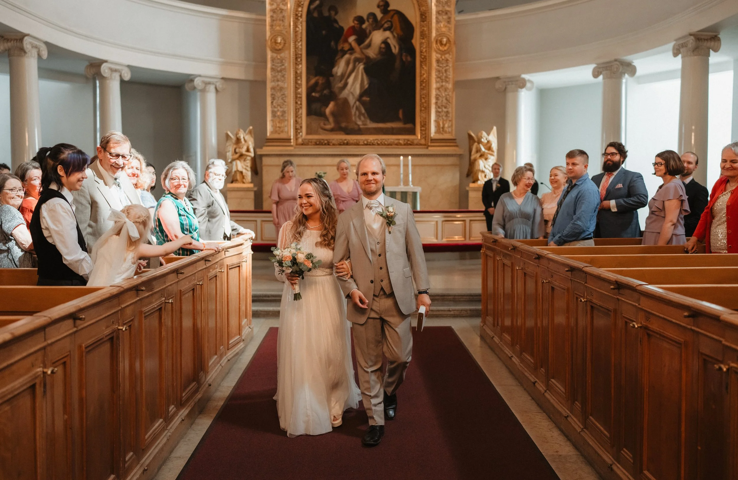 Bride and groom walking down the aisle in a church, surrounded by smiling guests, with an ornate painting in the background.