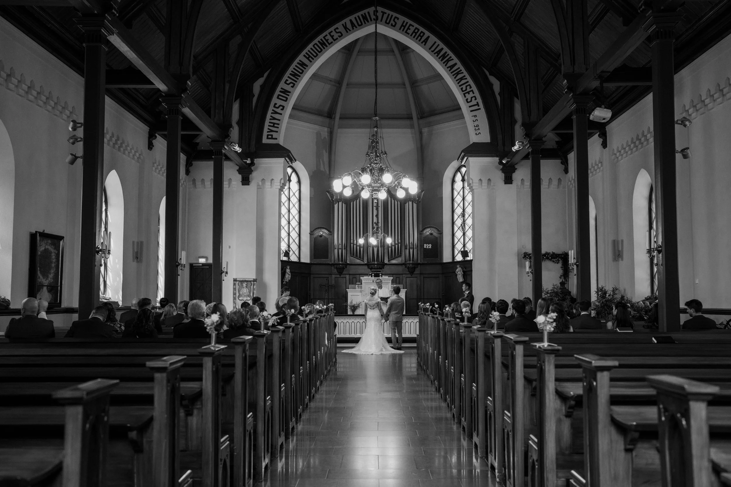 Black and white photo of a wedding ceremony in a church with a bride and groom standing at the altar, surrounded by guests seated in pews.