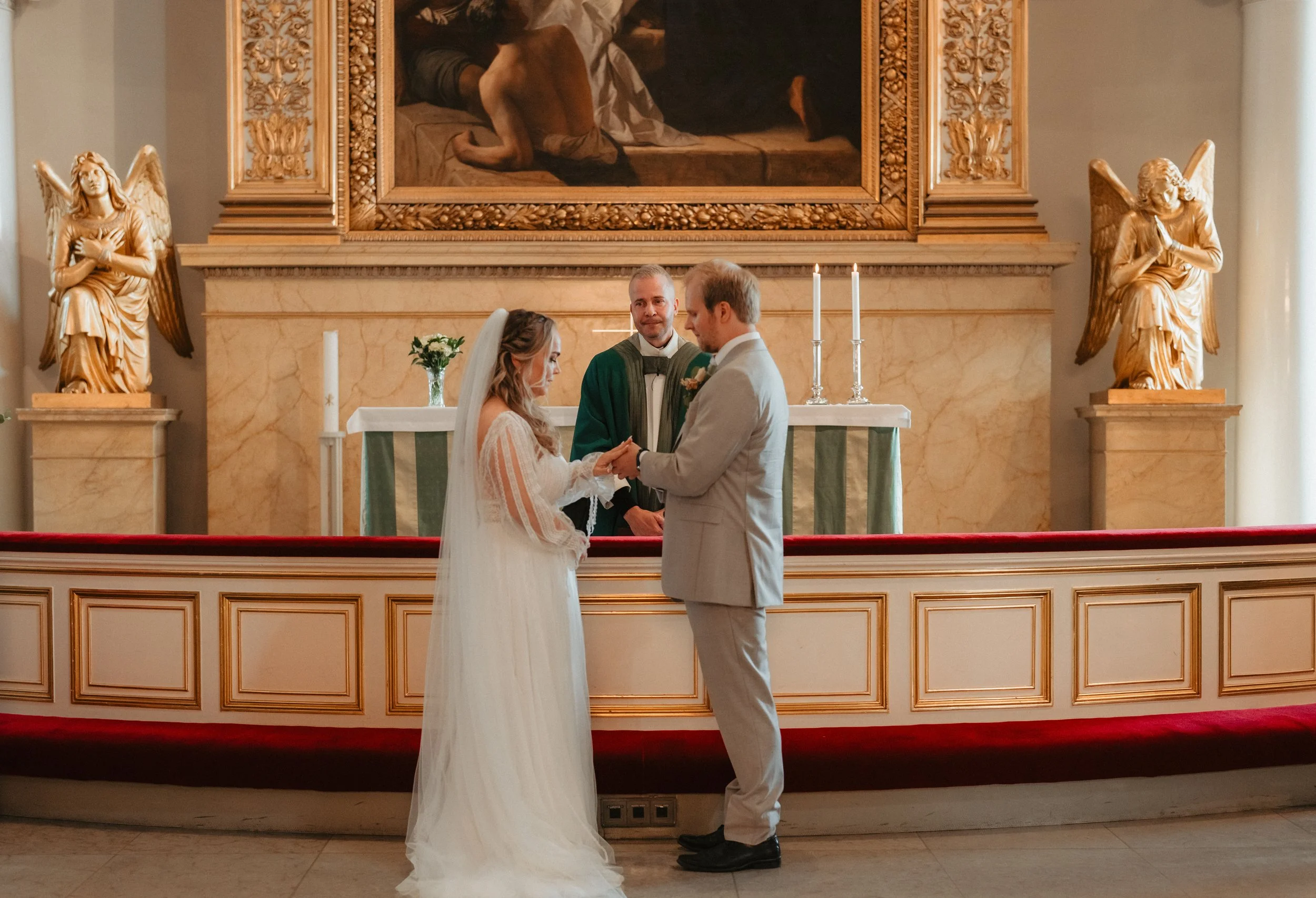 Wedding ceremony in a church with a bride and groom exchanging vows, officiant in the background, gold angel statues, and altar.