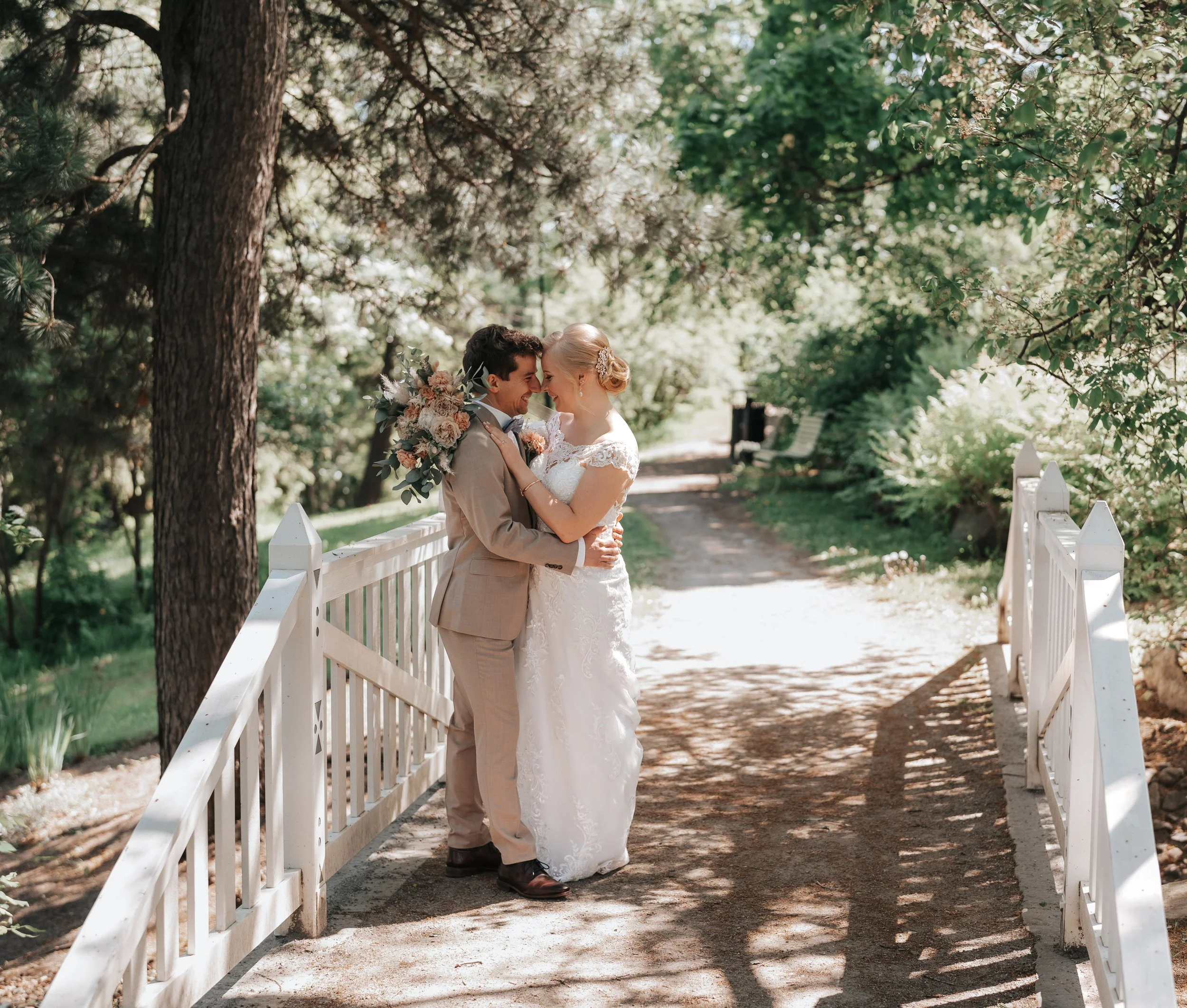 Bride and groom embracing on a white bridge surrounded by trees in a garden setting.
