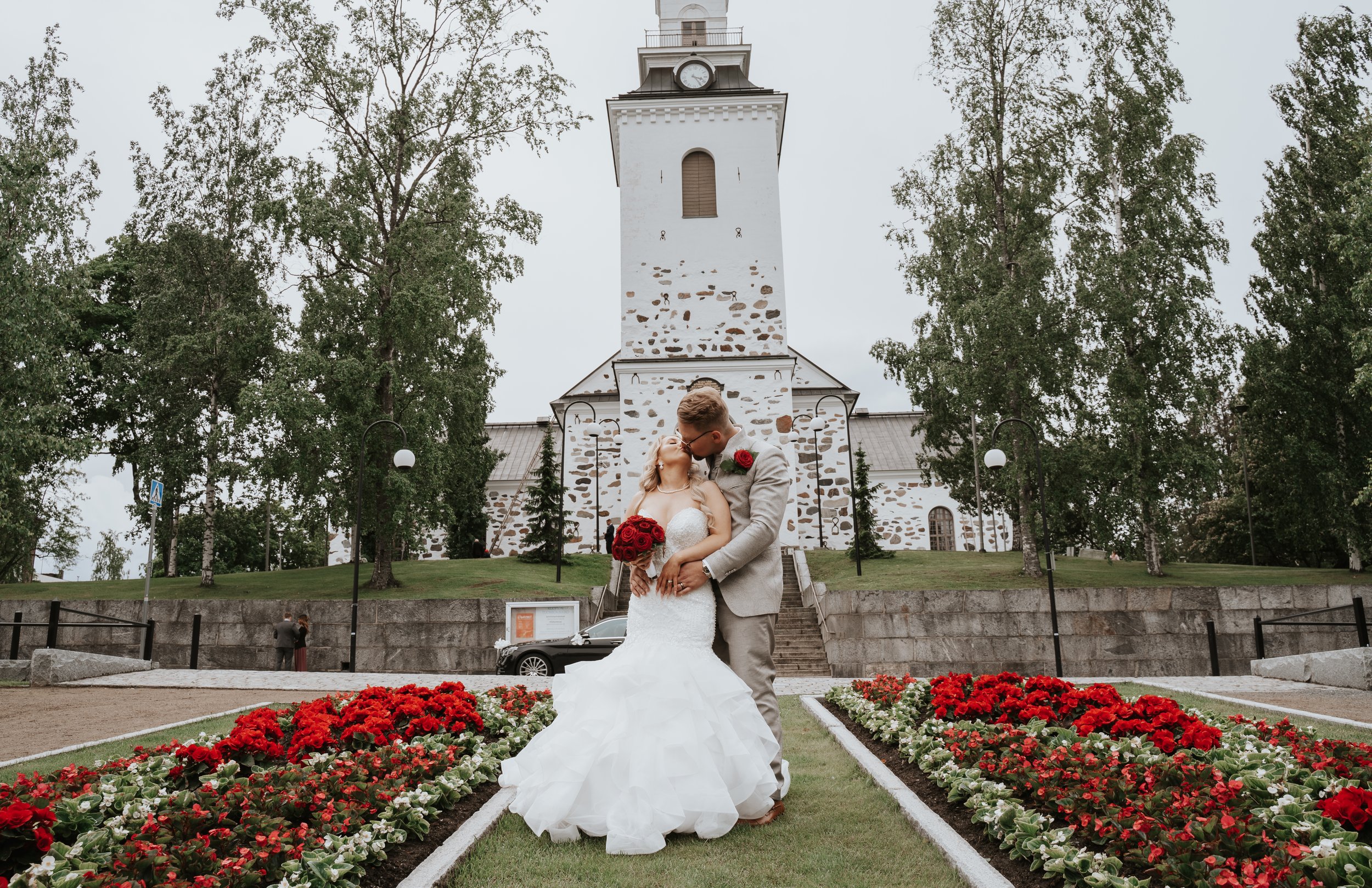 Wedding couple in front of a church, surrounded by trees and red flower beds