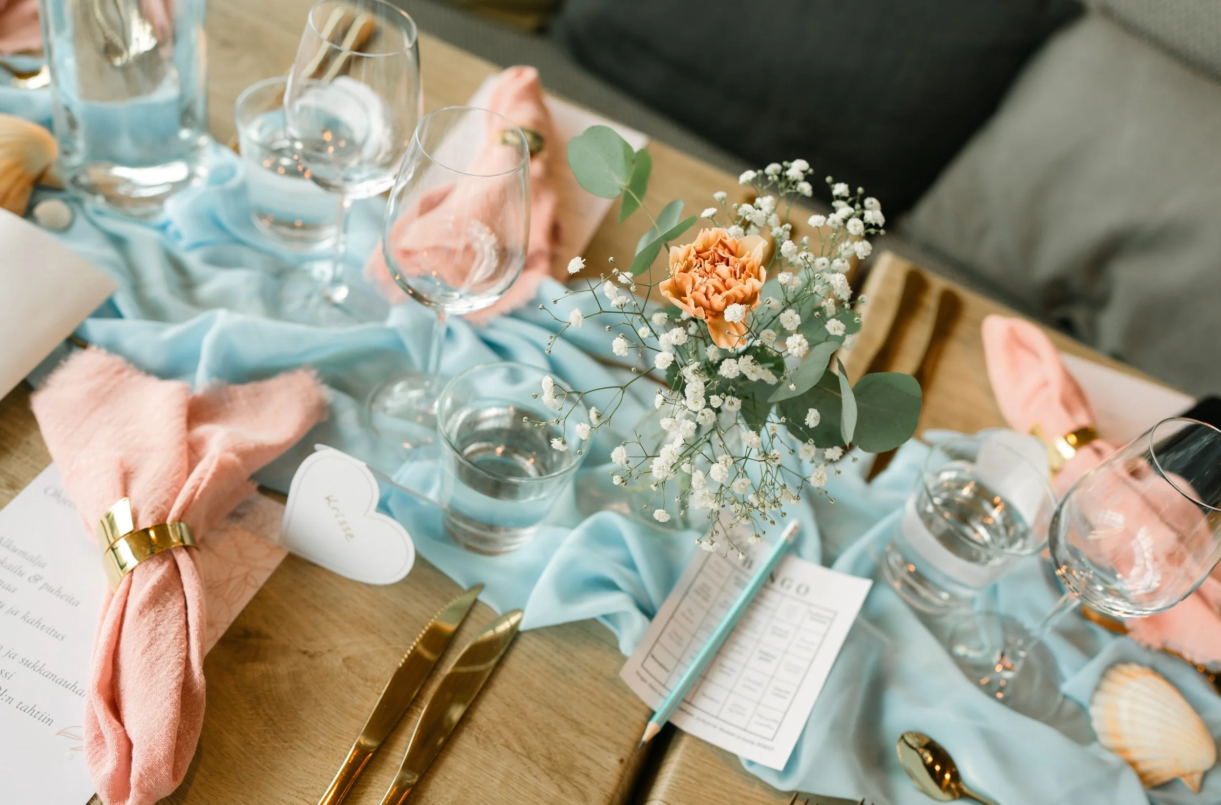 Elegant table setting with pink napkins, gold cutlery, crystal glasses, a flower centerpiece with orange and white flowers, and a blue table runner.