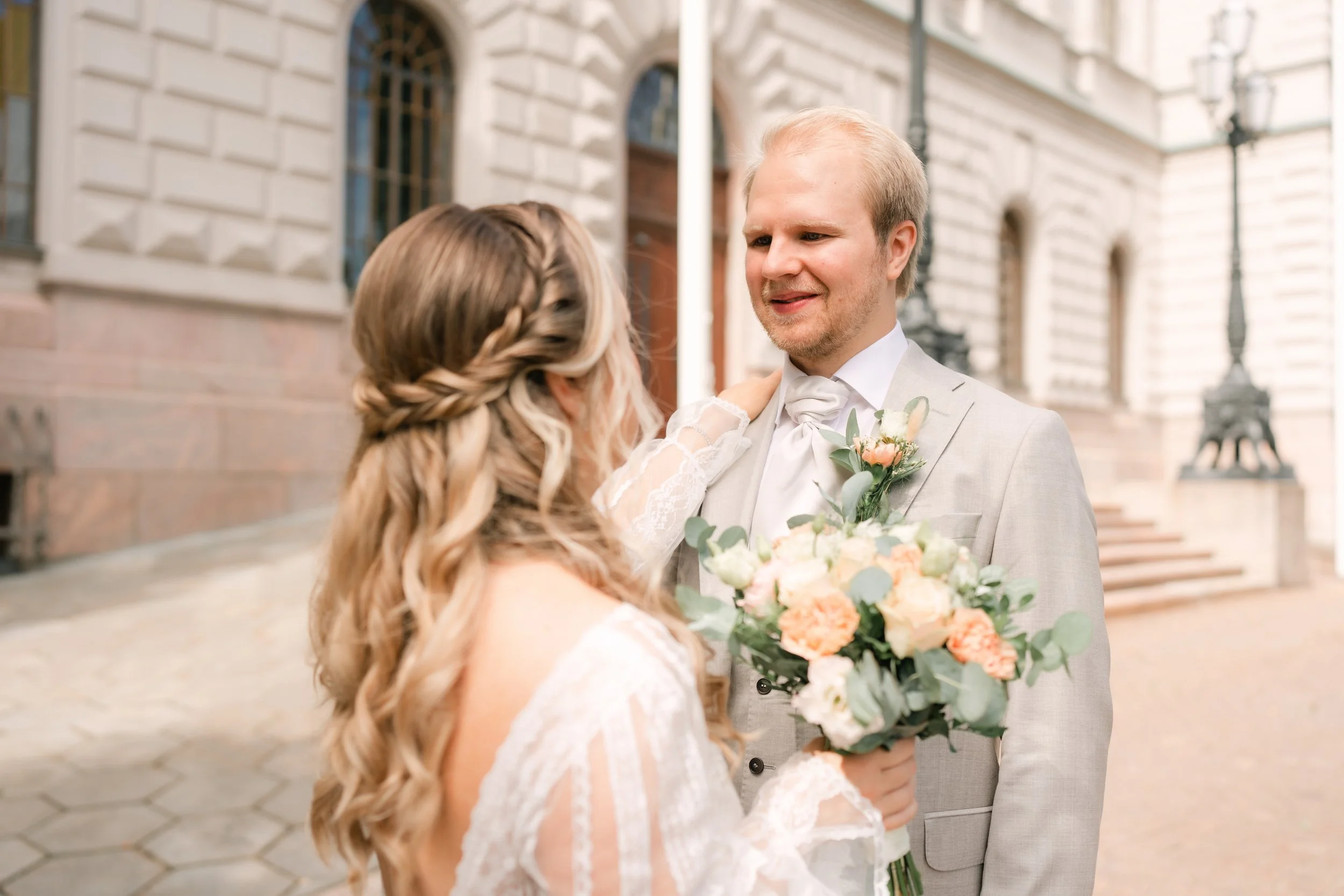 Bride and groom smiling in wedding attire, holding flowers, outside a building.