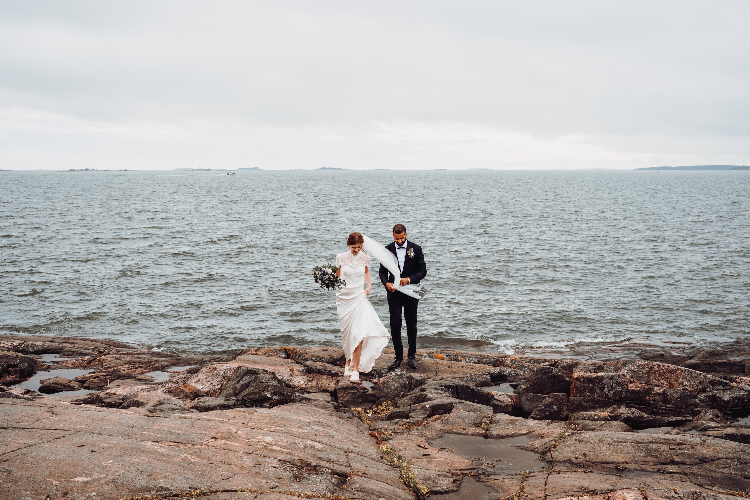 Bride and groom walking on rocky shore by the sea, wedding attire, overcast sky.