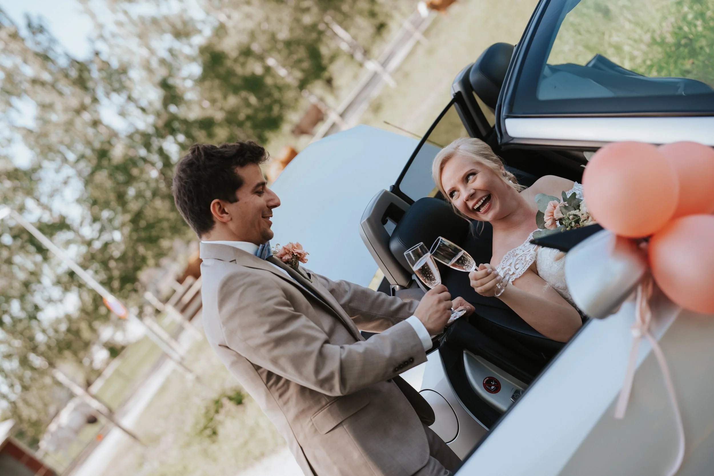 Bride and groom toasting with champagne glasses in a convertible decorated with balloons.