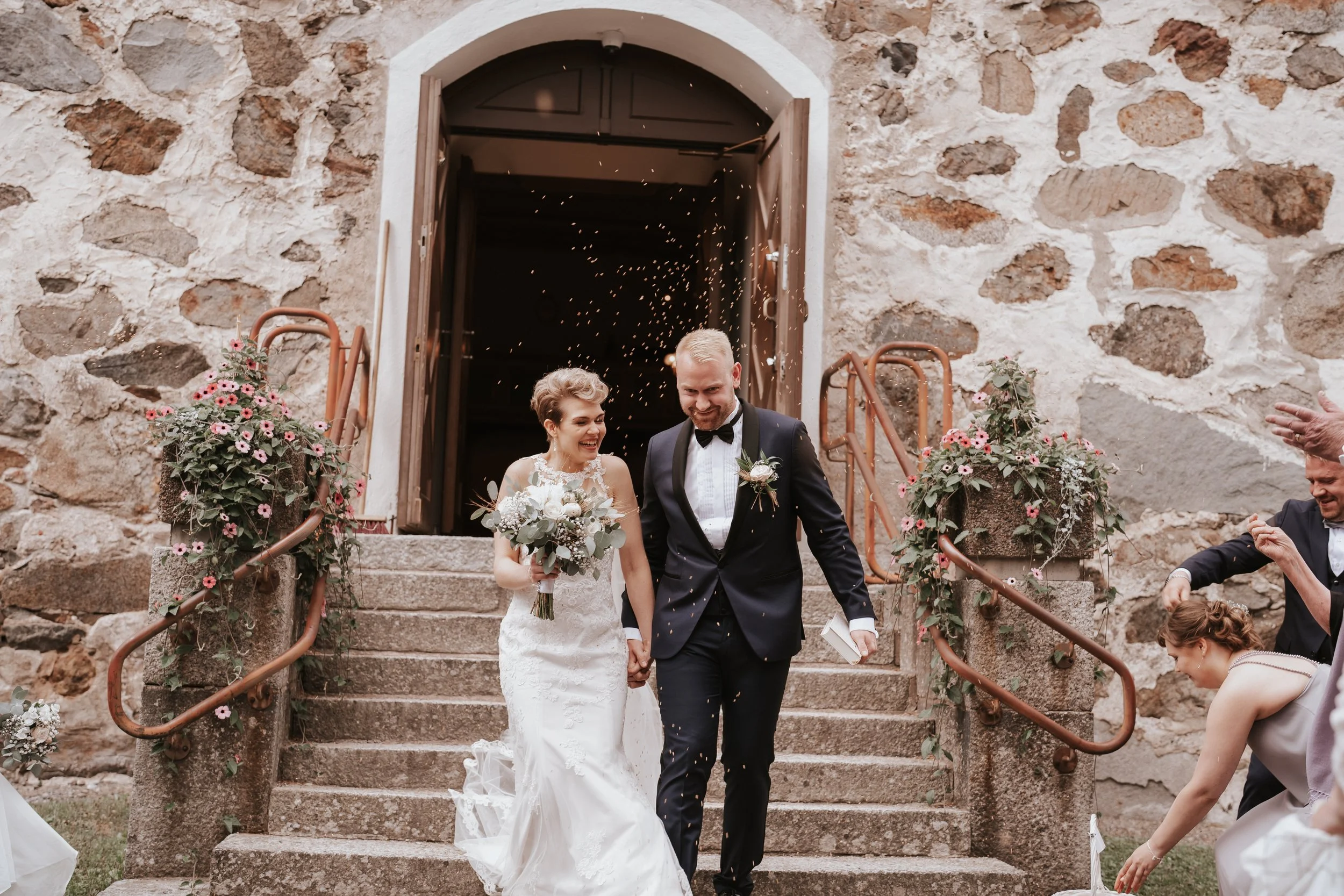 Bride and groom walking down church steps, smiling, with flower bouquets and confetti, surrounded by wedding guests and floral decorations.