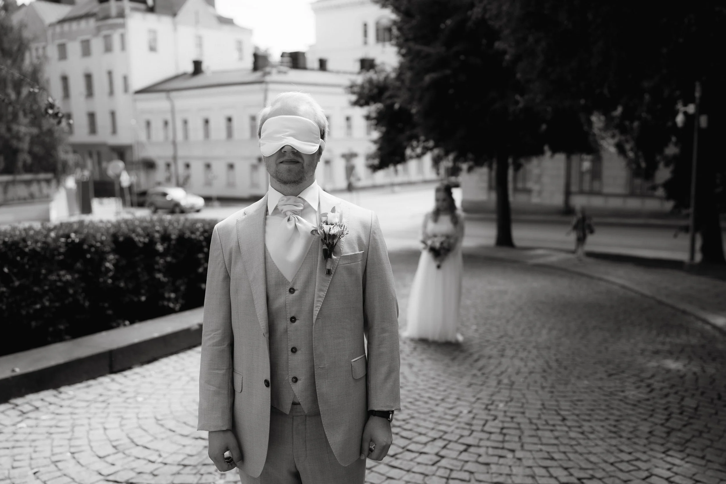 Blindfolded groom in suit with boutonnière standing outdoors in front of blurred bride holding bouquet.