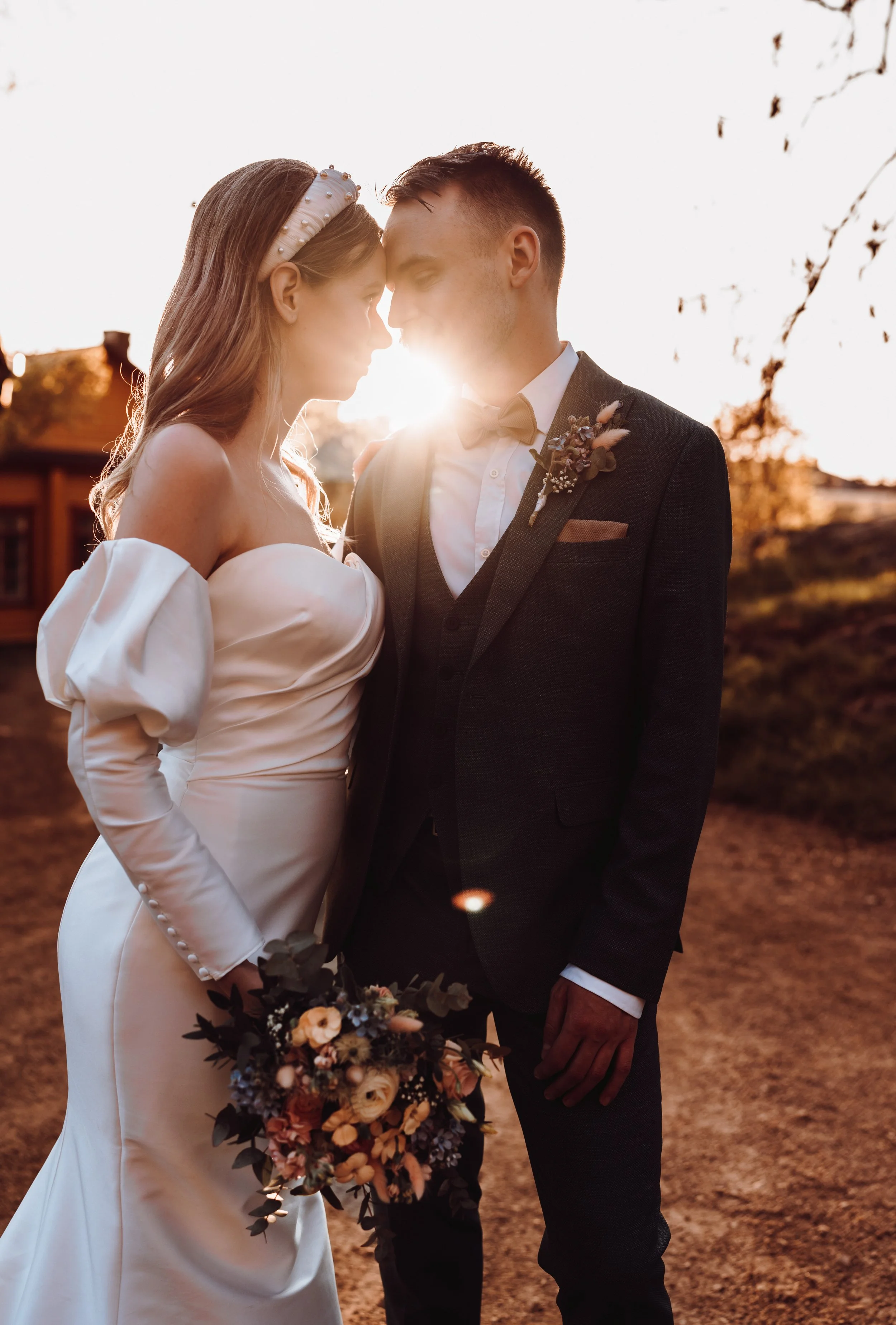 Bride and groom standing close, holding bouquet, sunlight in background, outdoor setting.