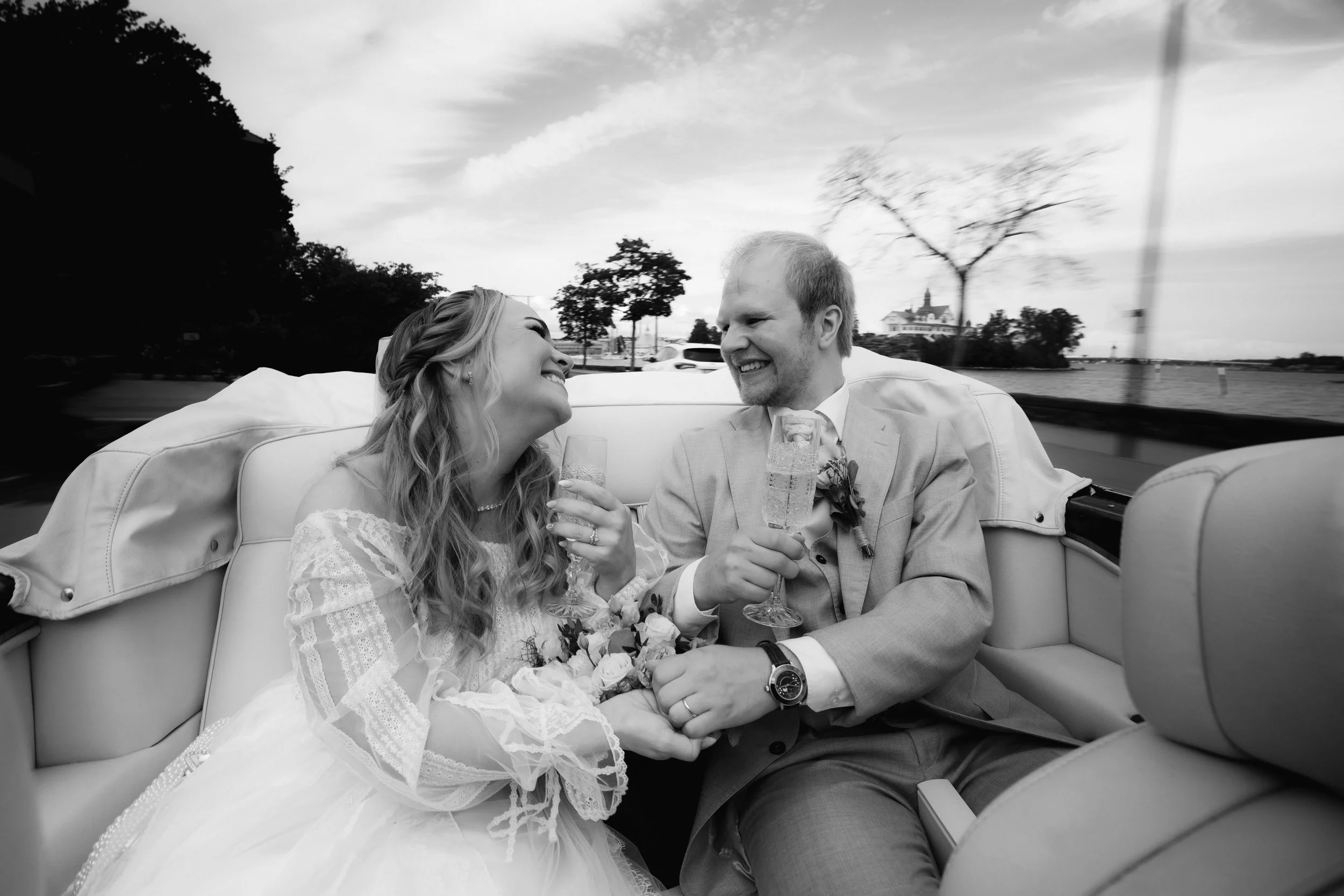 Bride and groom laughing while holding champagne glasses in a convertible.