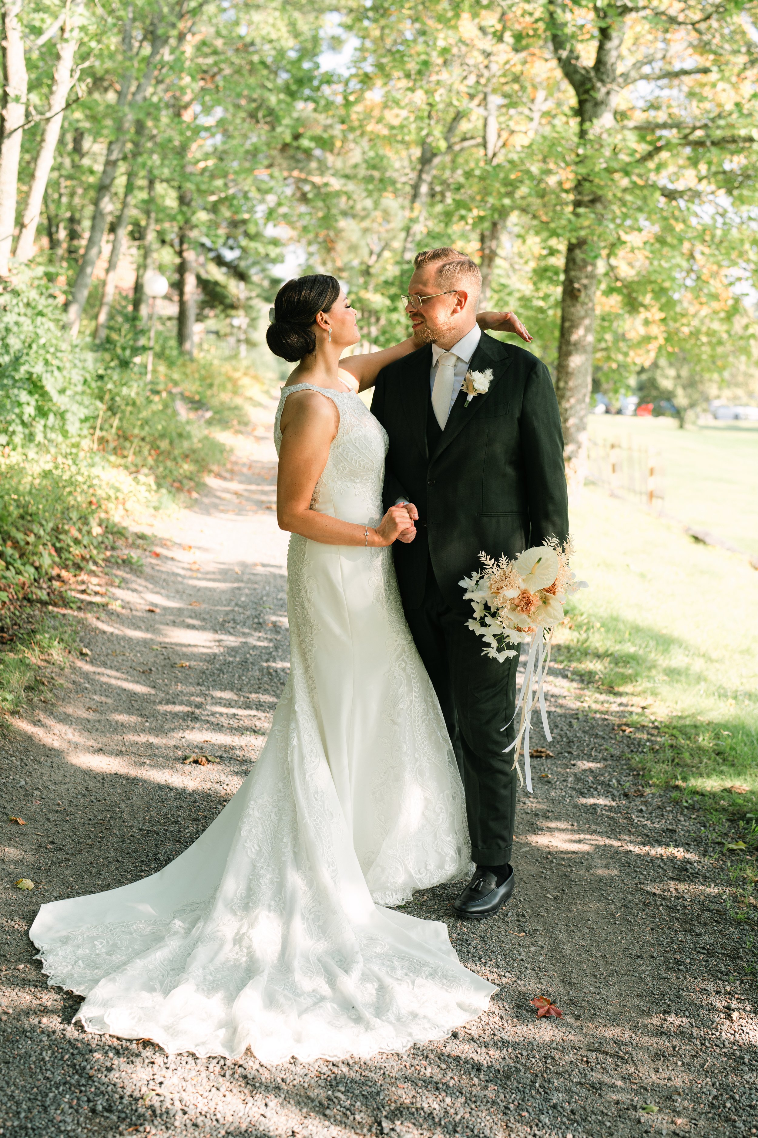 Bride and groom on a forested path, bride holding bouquet, wearing a white wedding gown, groom in black suit, surrounded by greenery.