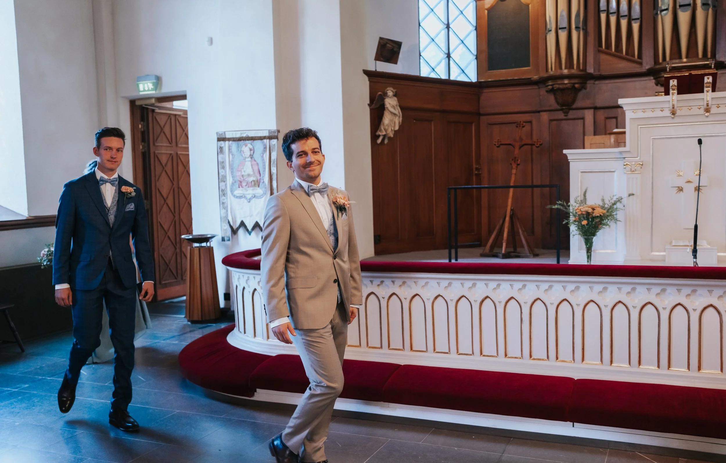 Two men in suits walking down the aisle of a church, decorated with a white altar and floral arrangements, during a wedding ceremony.