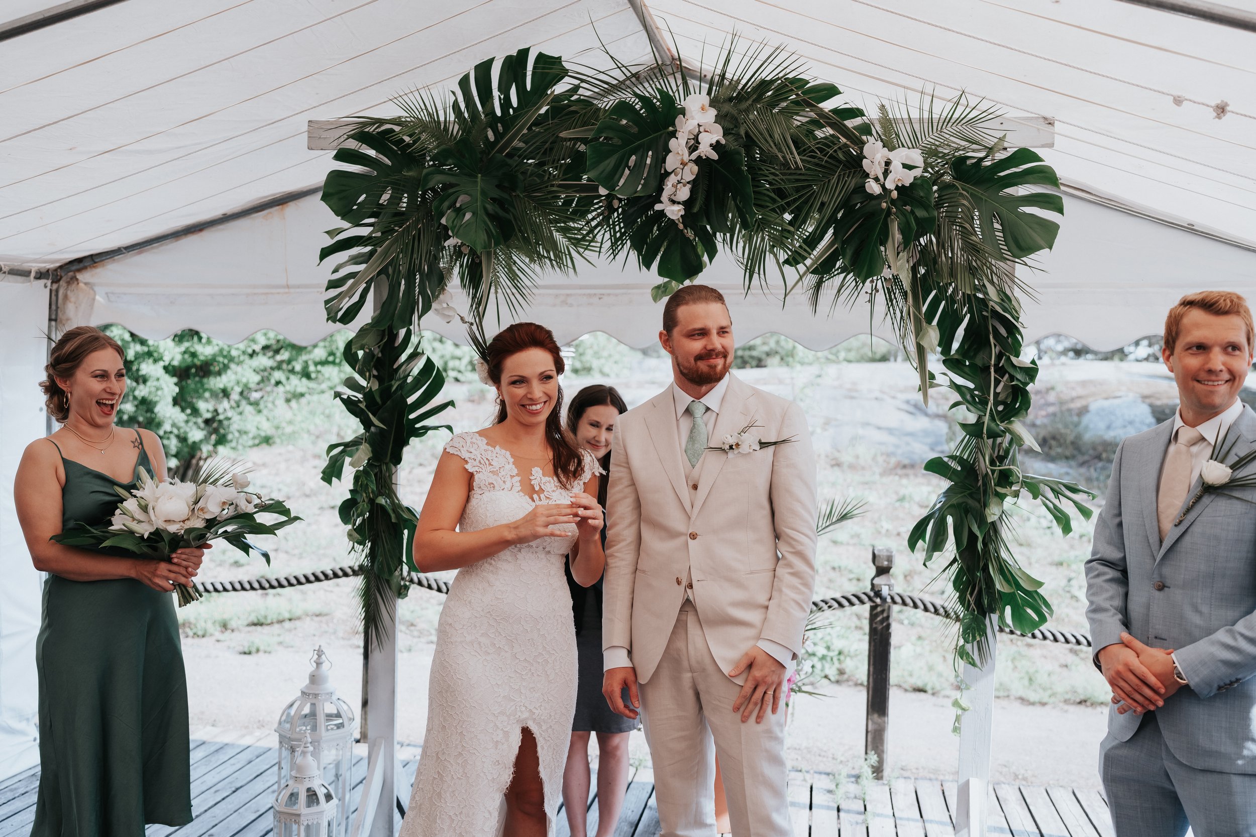 Wedding ceremony under a tent with tropical greenery and floral arch; bride in white dress, groom in beige suit, bridesmaid holding bouquet.
