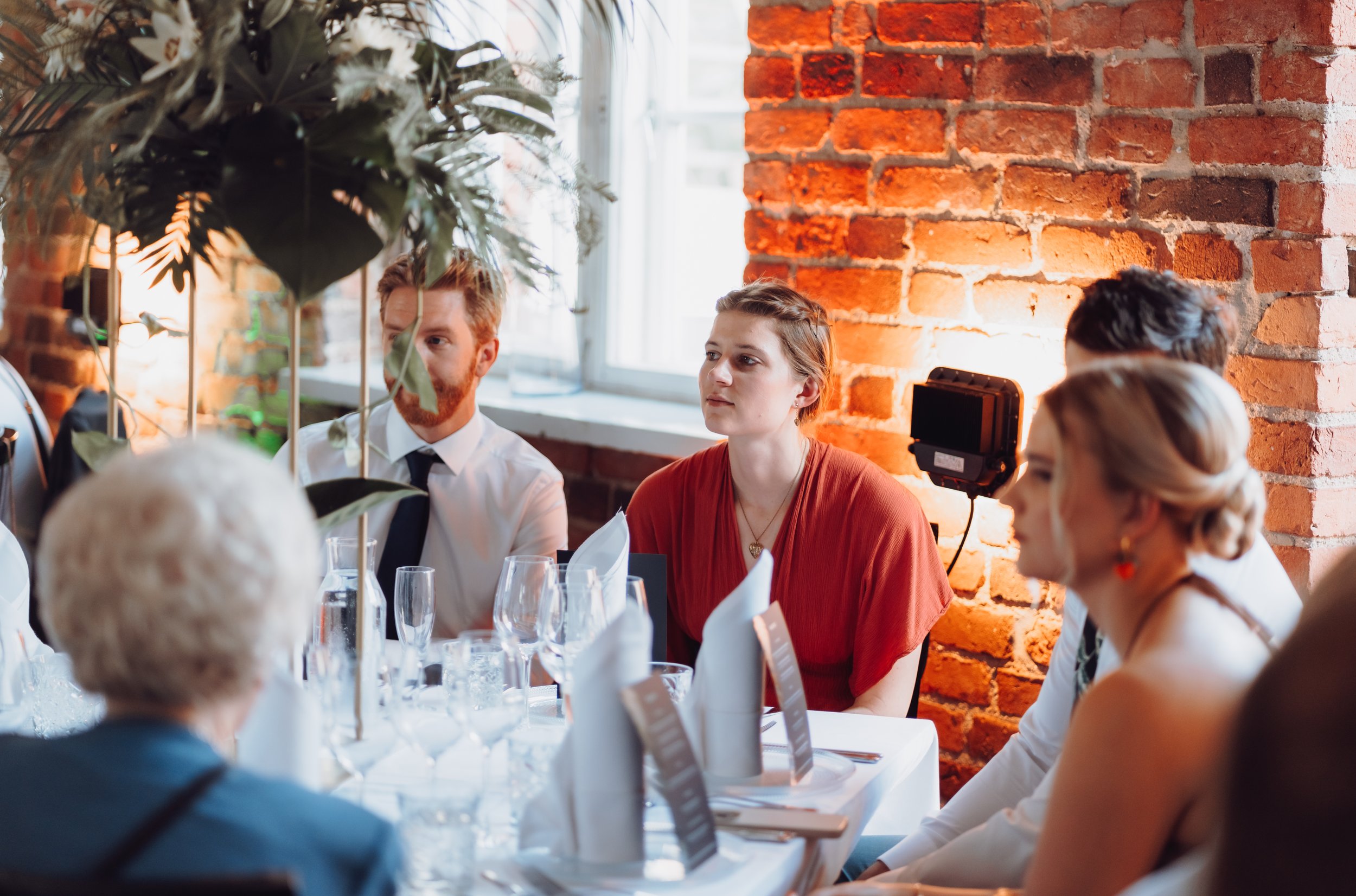 Group of people sitting at a table set for a formal meal, with floral centerpiece and exposed brick wall in the background.
