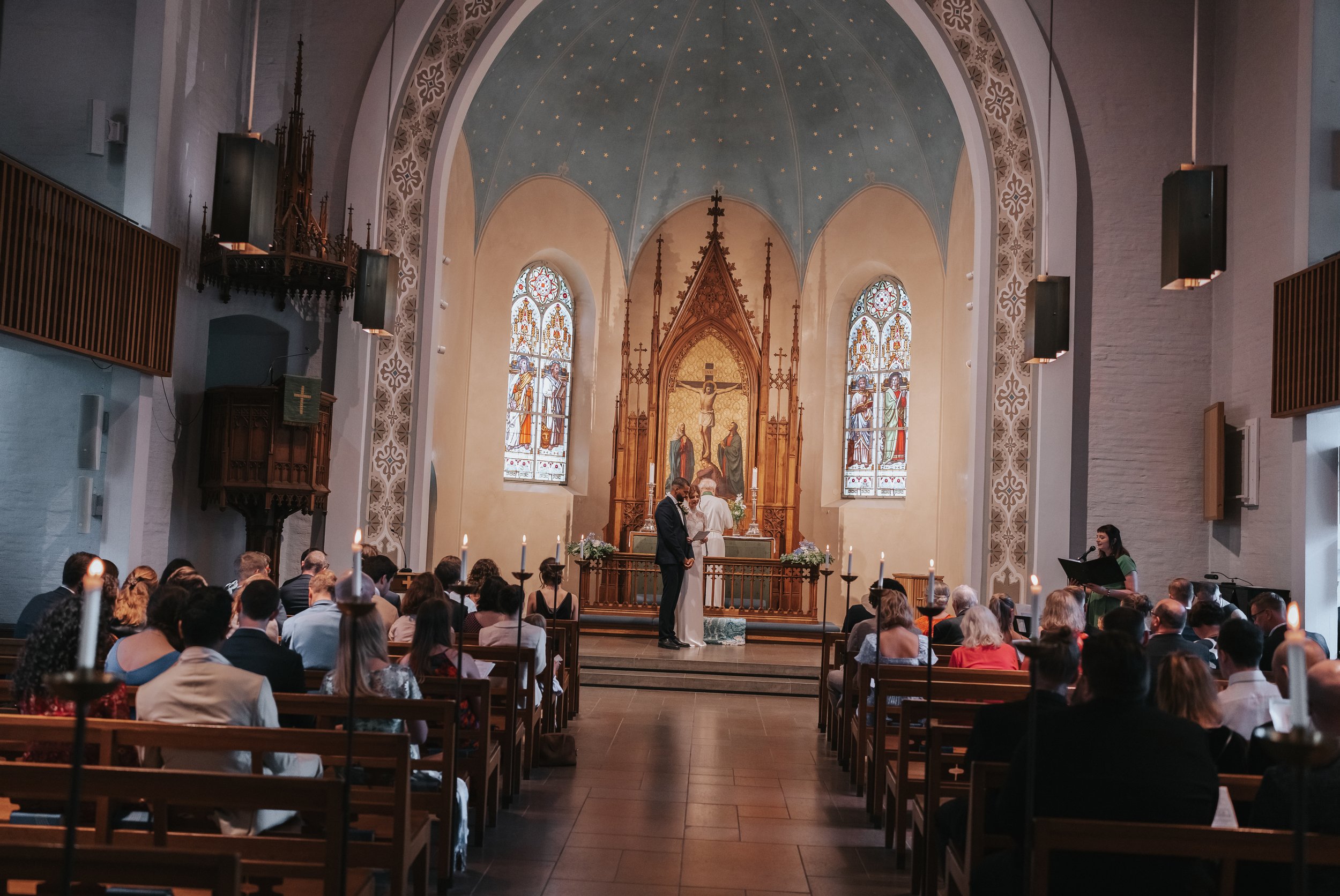 Wedding ceremony in a church with stained glass windows and an ornate altar, bride and groom at the front and seated guests observing.