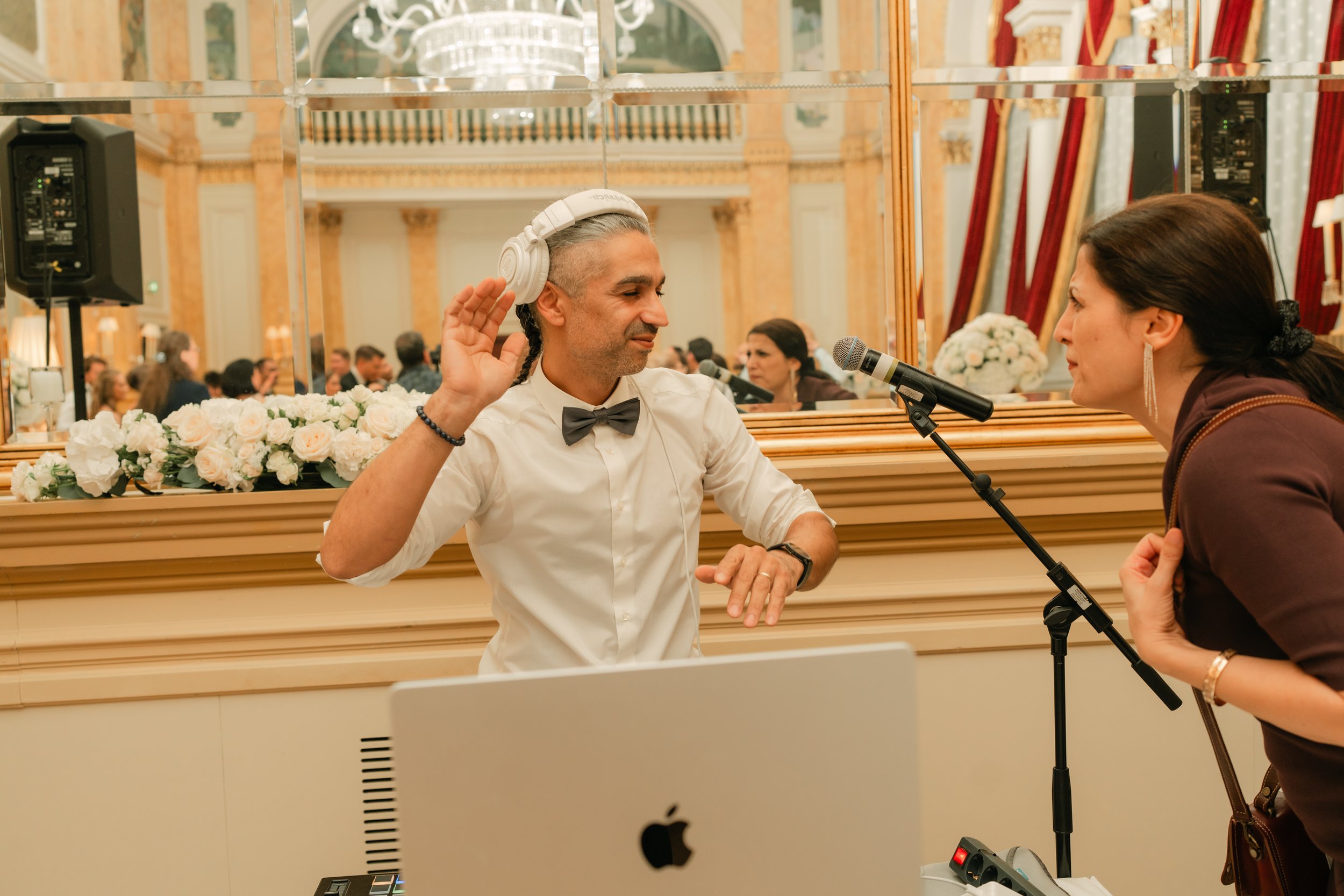 DJ with headphones at an event, talking to a woman with a microphone, elegant ballroom setting, chandelier, floral arrangement, ornate decor.