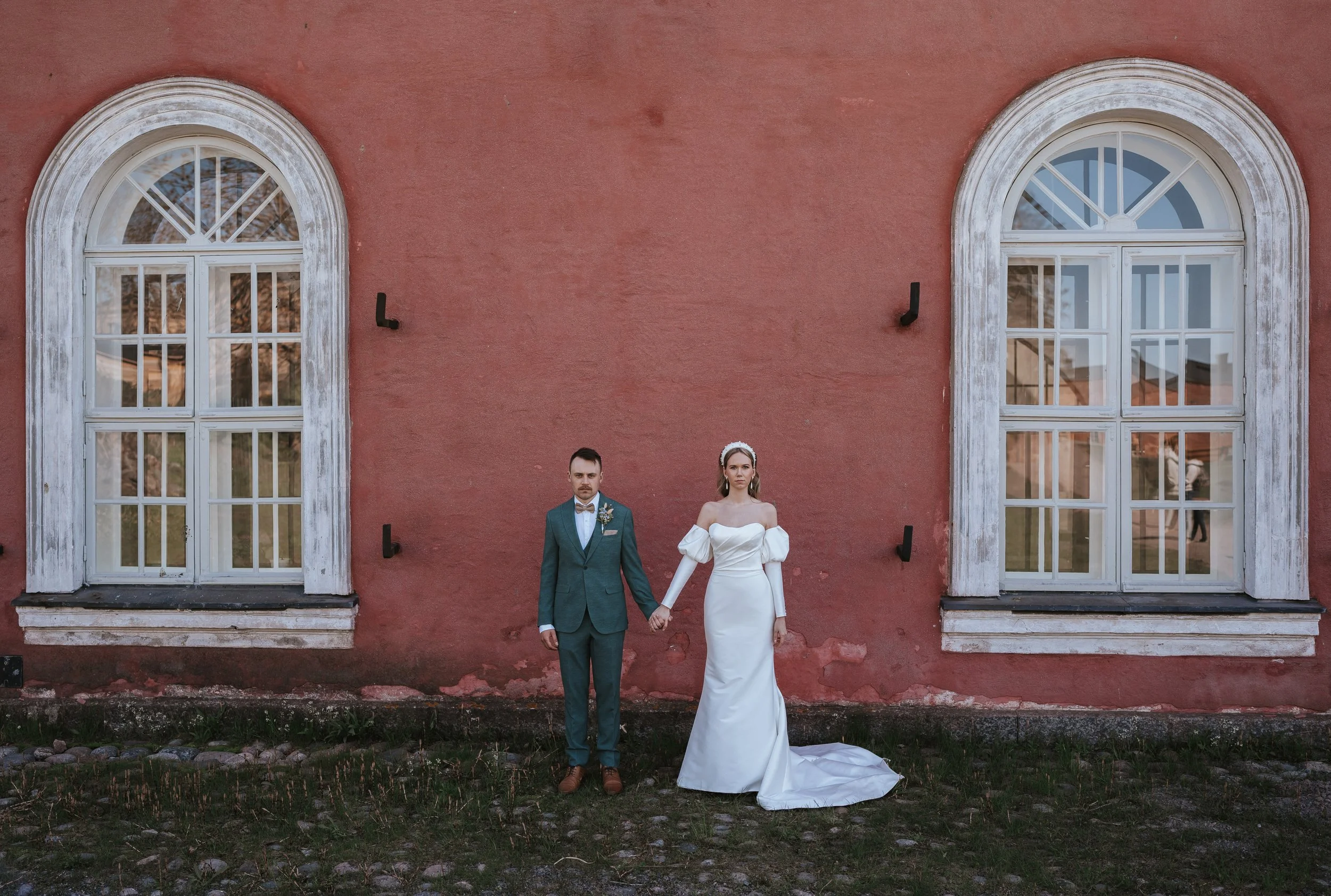 Bride and groom standing against a red wall with arched windows, holding hands.