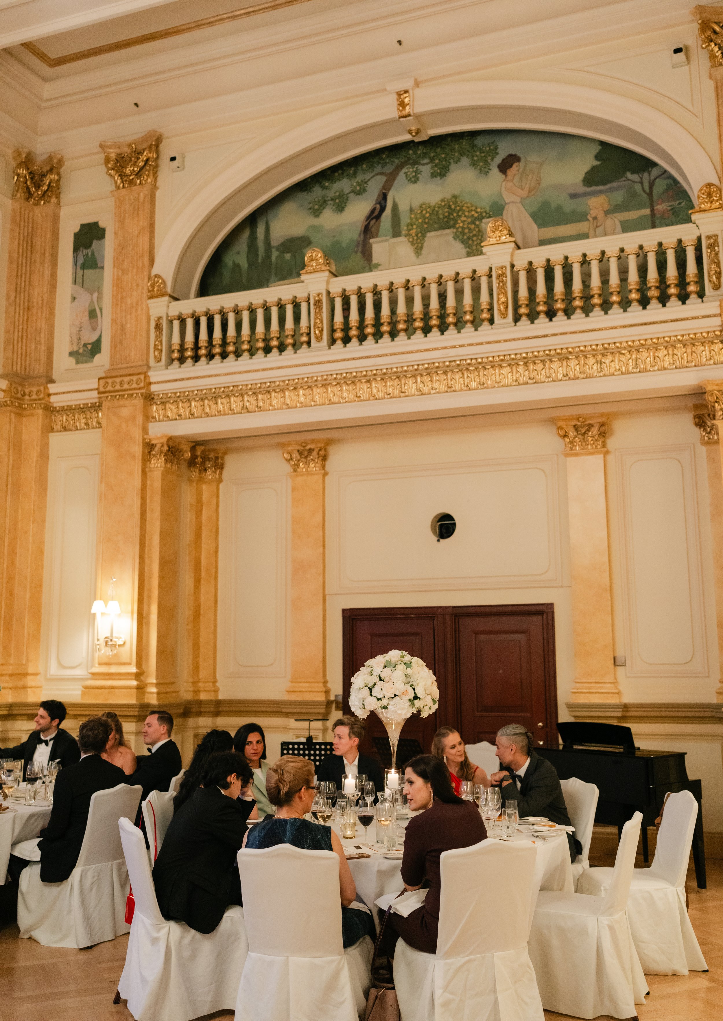 Elegant formal dining event in a grand hall with ornate architecture, featuring guests seated at round tables, adorned with floral centerpieces, under intricate wall murals and a decorative balcony.