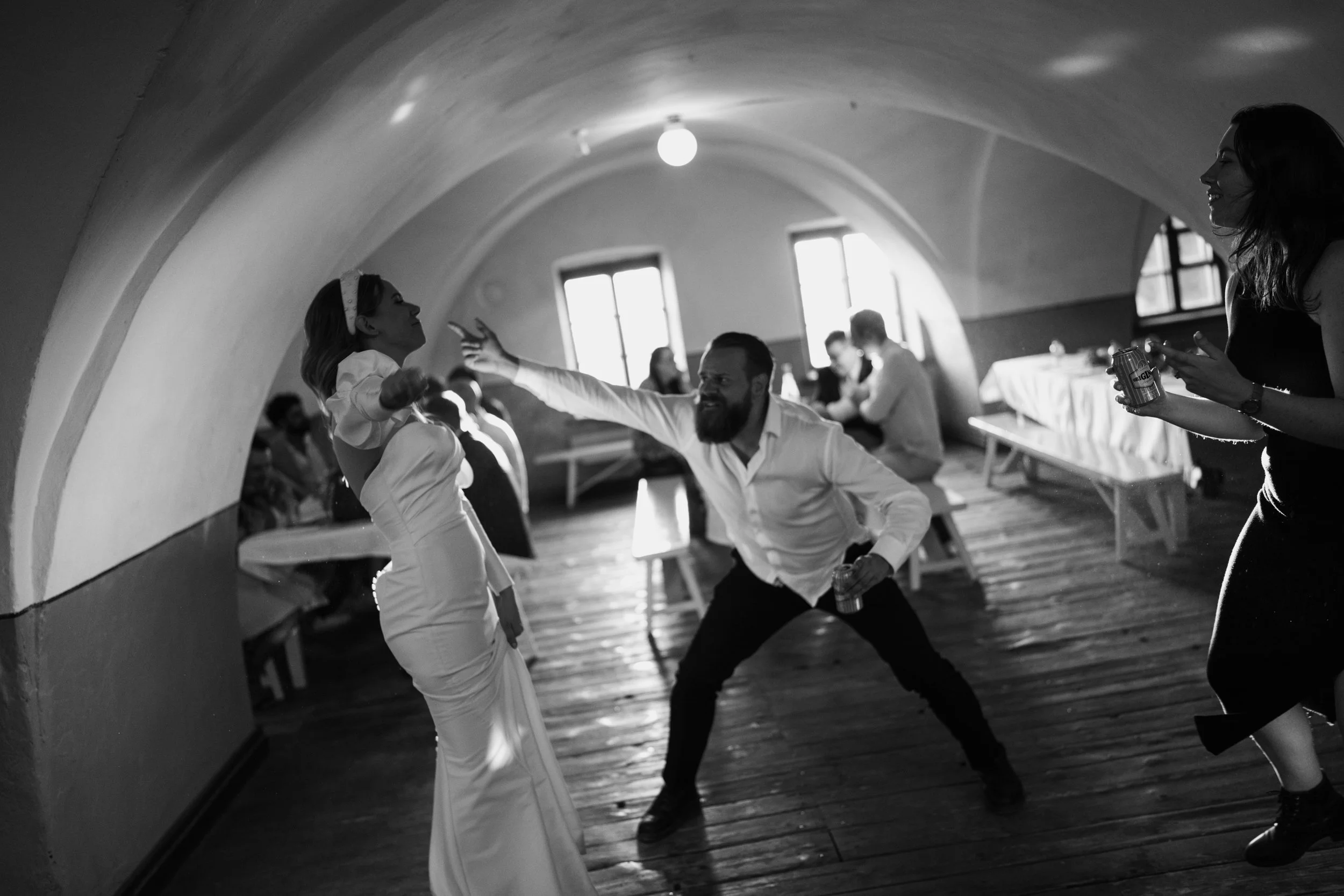 Black and white photo of people dancing energetically in a room with arched ceilings, with a visible bride in a wedding dress on the left.
