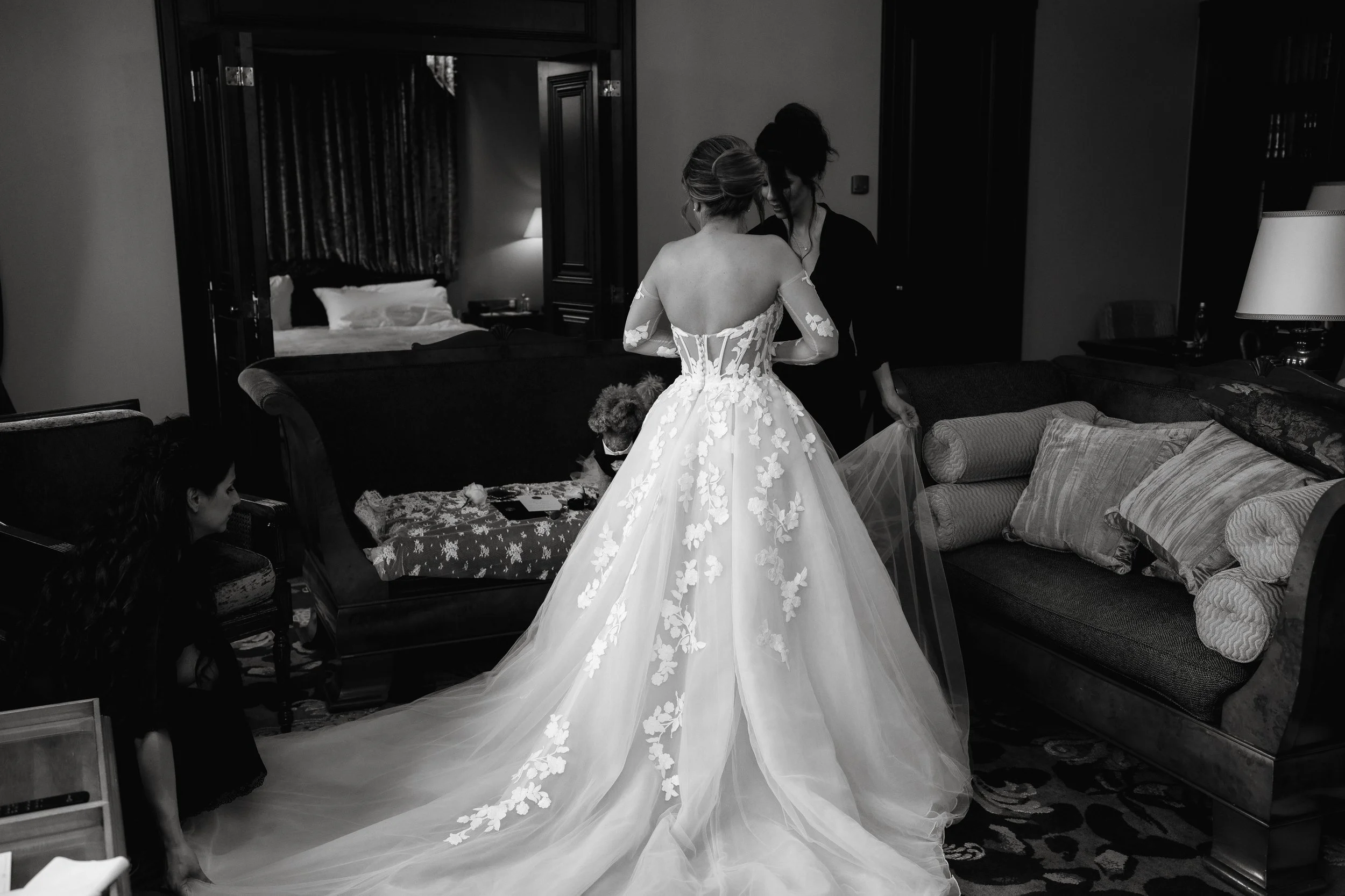 Black and white photo of a bride in a floral bridal gown being assisted by two people in a room with a sofa and cushions.