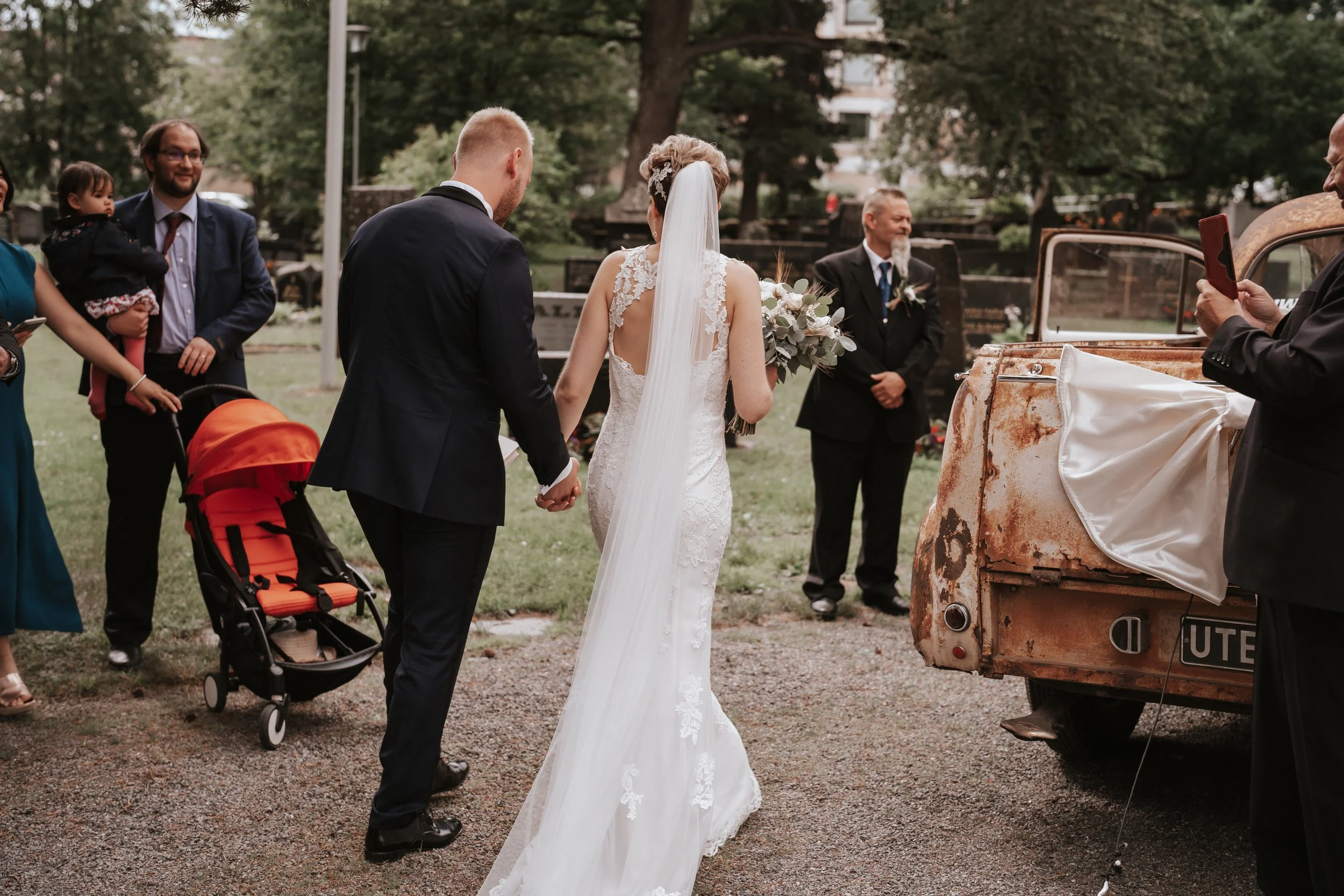 Wedding scene with bride in white dress holding flowers, groom in suit, guests, red stroller, rusted car, and greenery background.