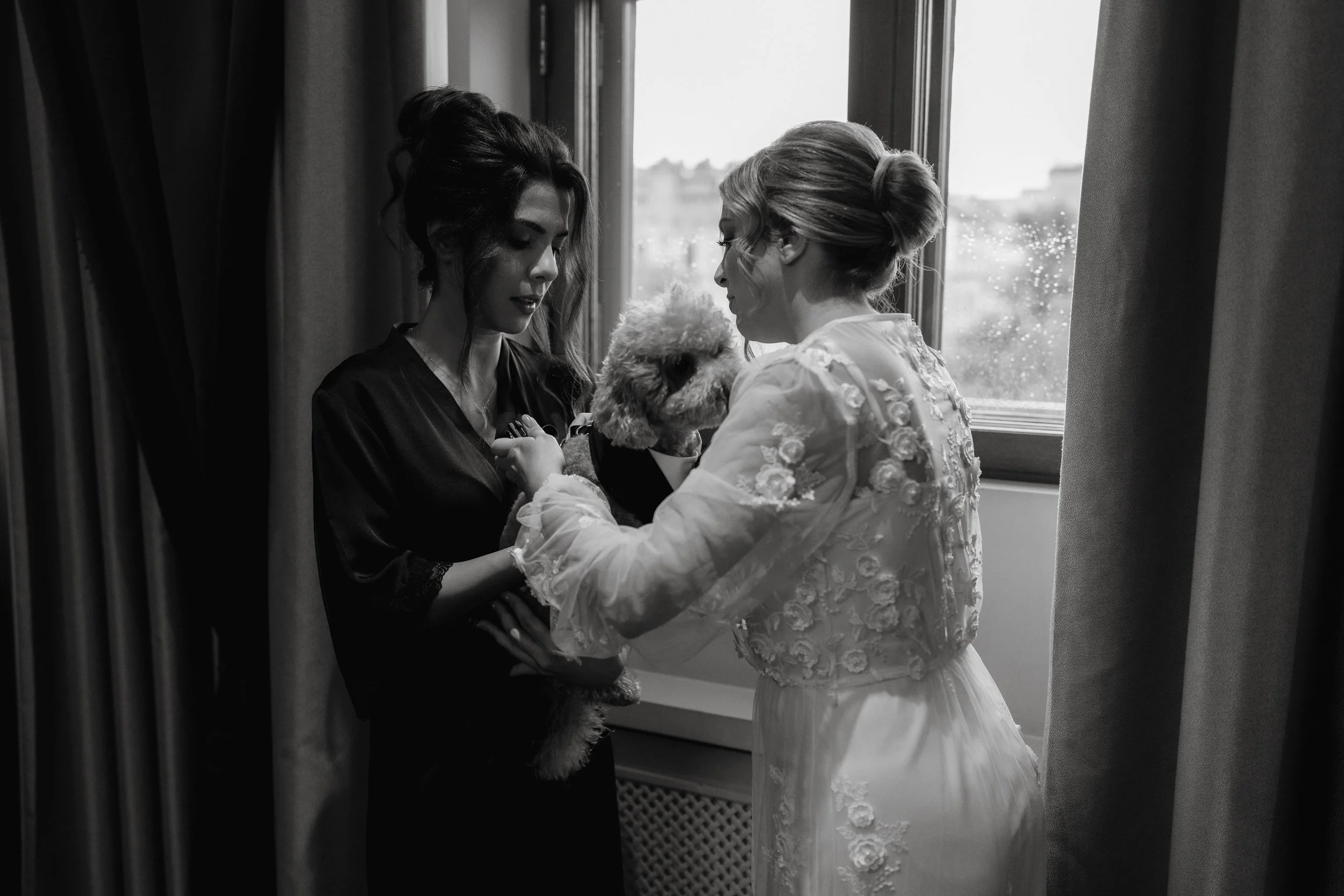 Black and white photo of two women in robes holding a dog near a window.