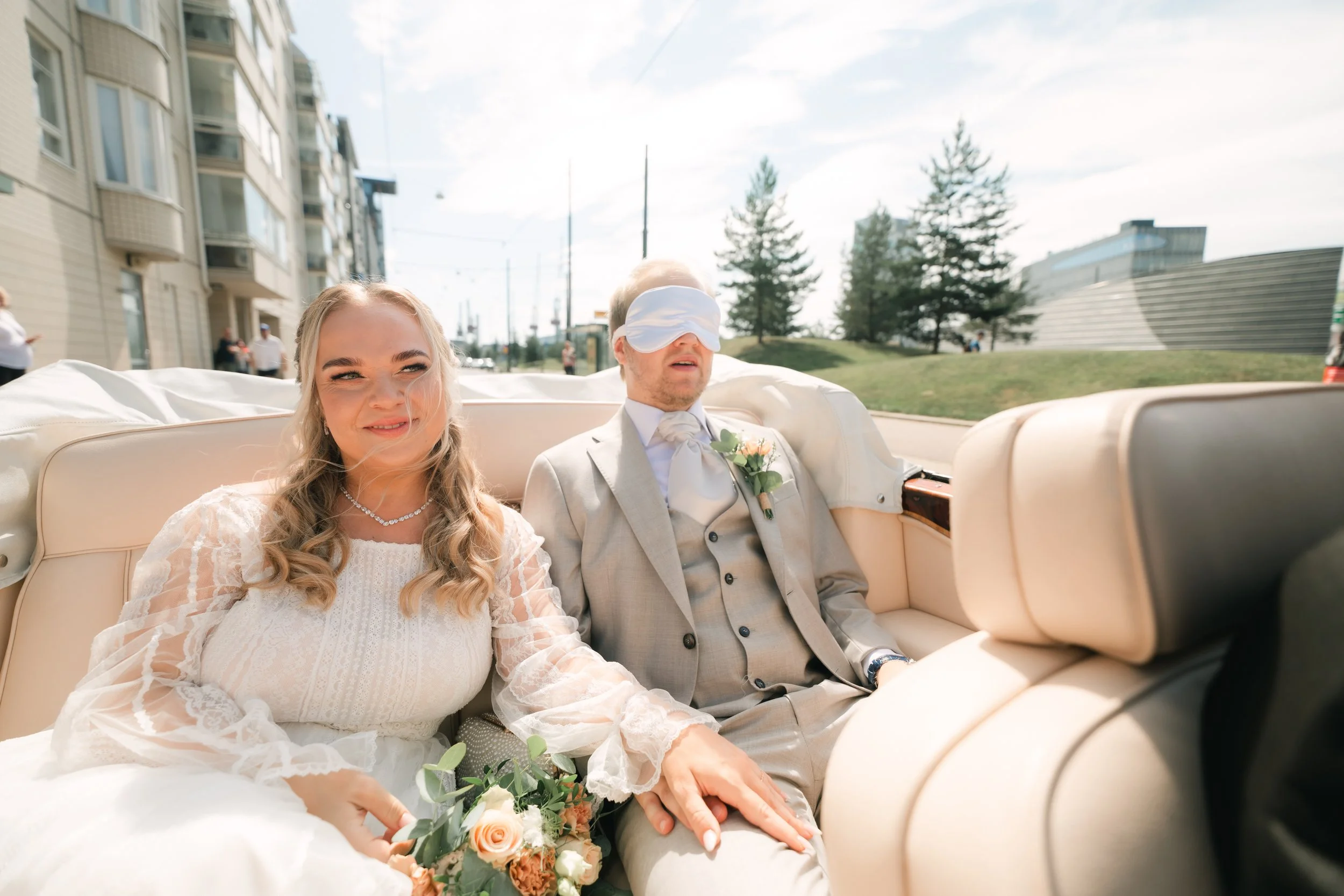 Bride and blindfolded groom in convertible on wedding day with buildings in background.