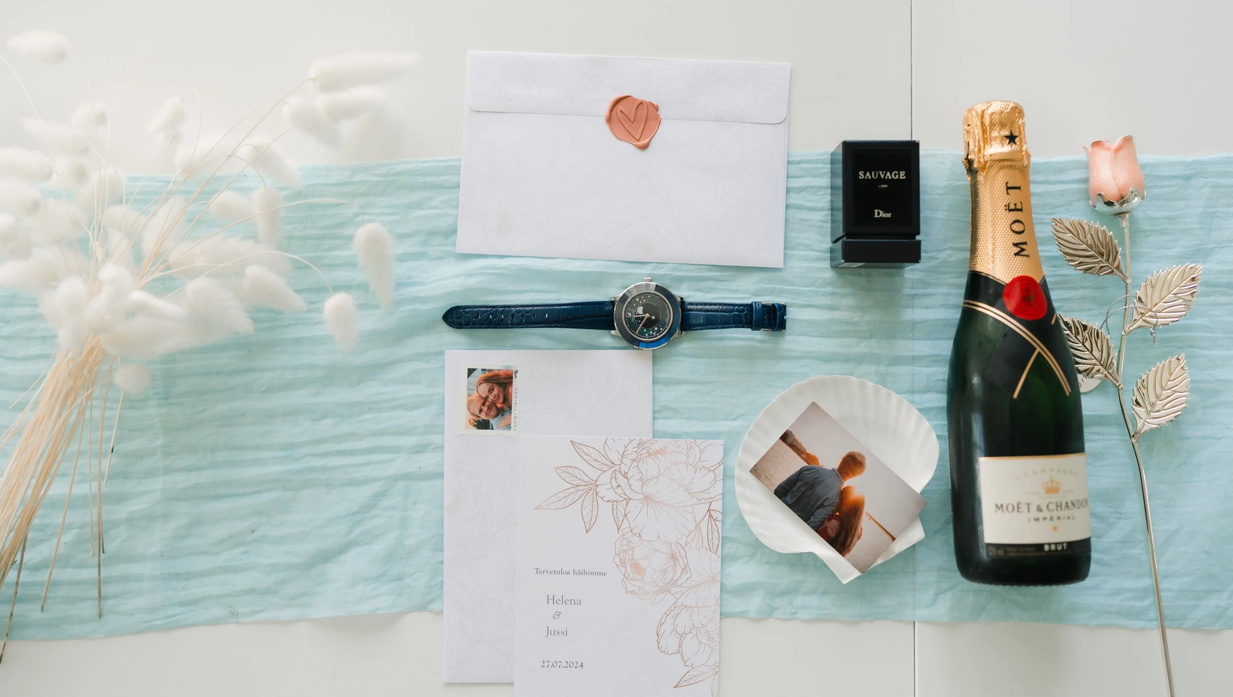 Flat lay of wedding invitation set with envelope, watch, perfume, champagne bottle, dried flowers, and a photo on a blue cloth background.