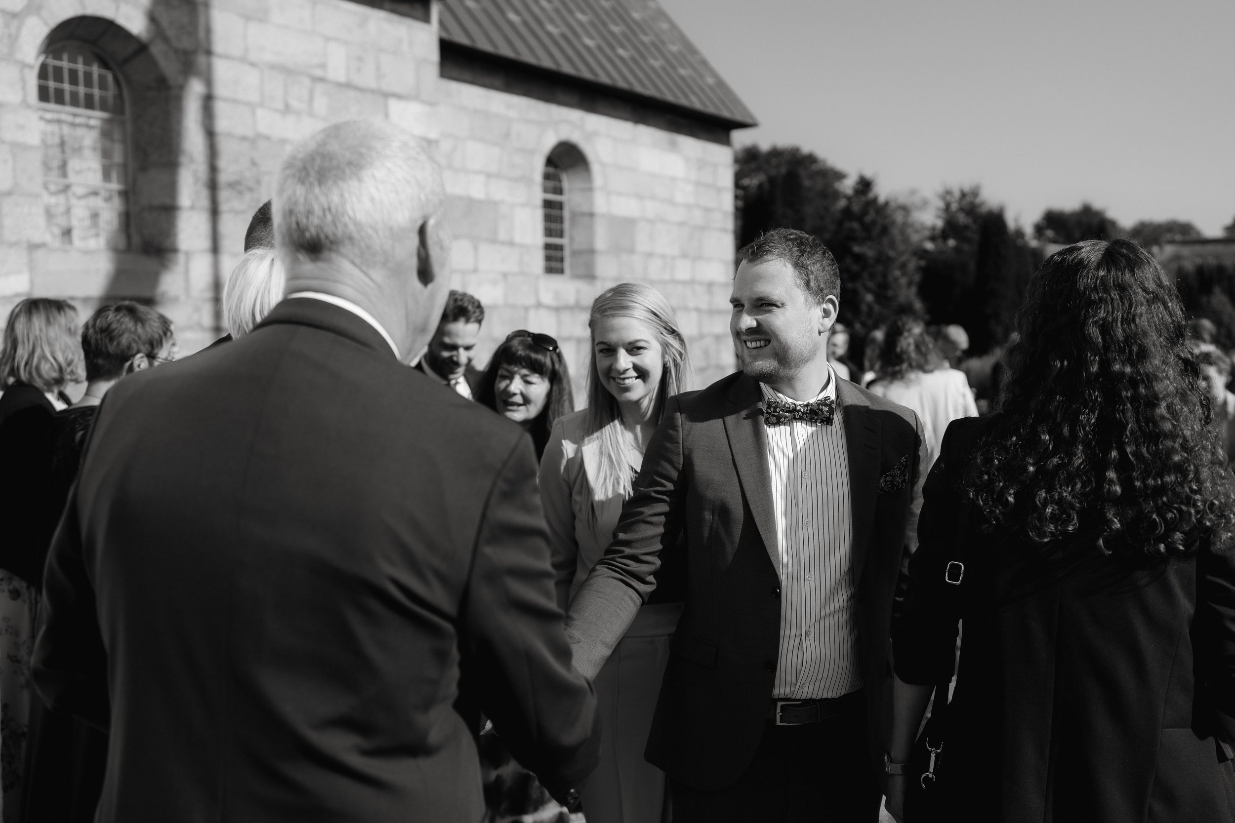 Group of people in formal attire shaking hands at an outdoor event, in front of a stone building.