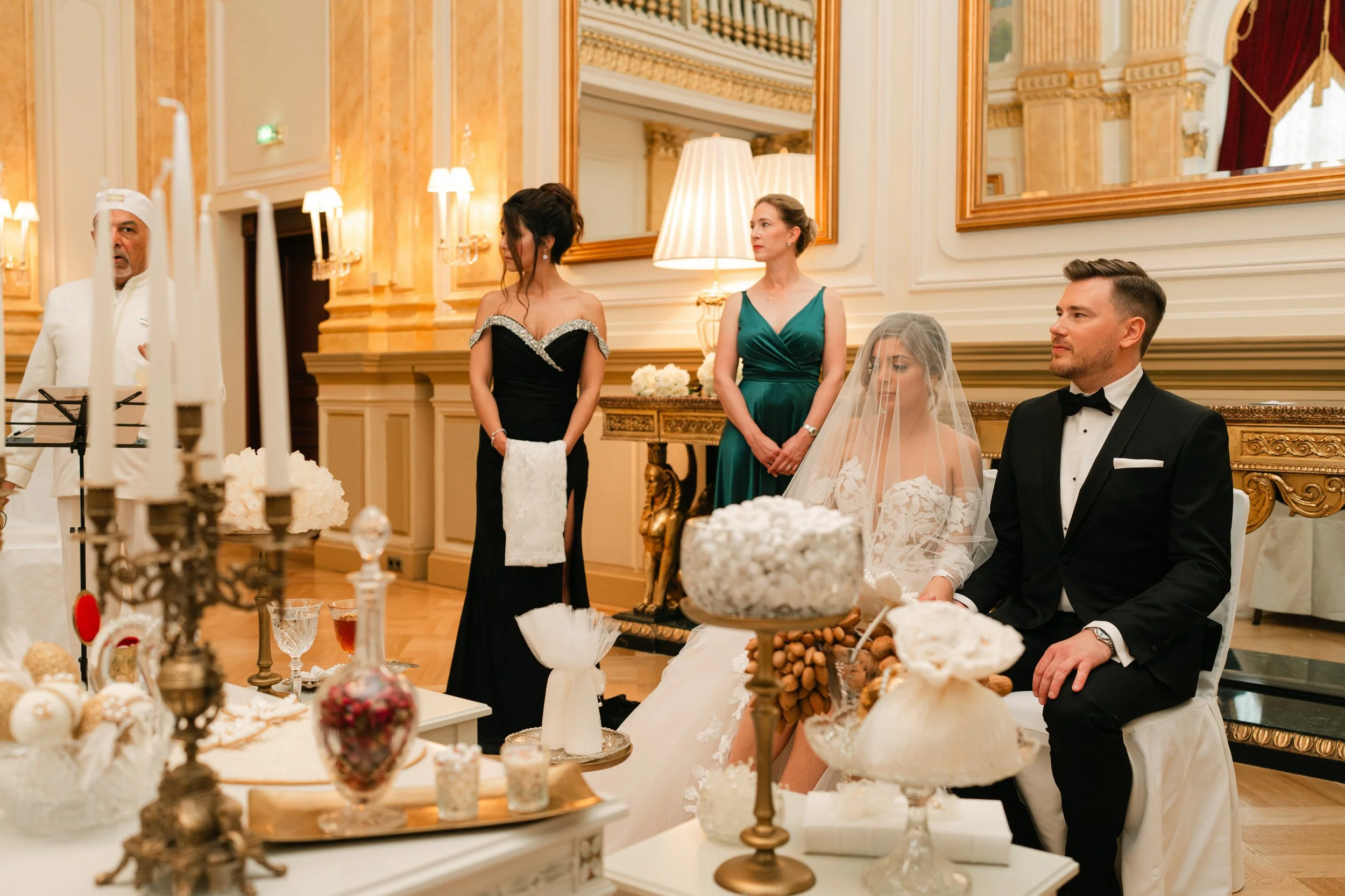 Wedding ceremony with bride and groom sitting, surrounded by decorative items, and accompanied by attendants in formal attire in an elegant room.