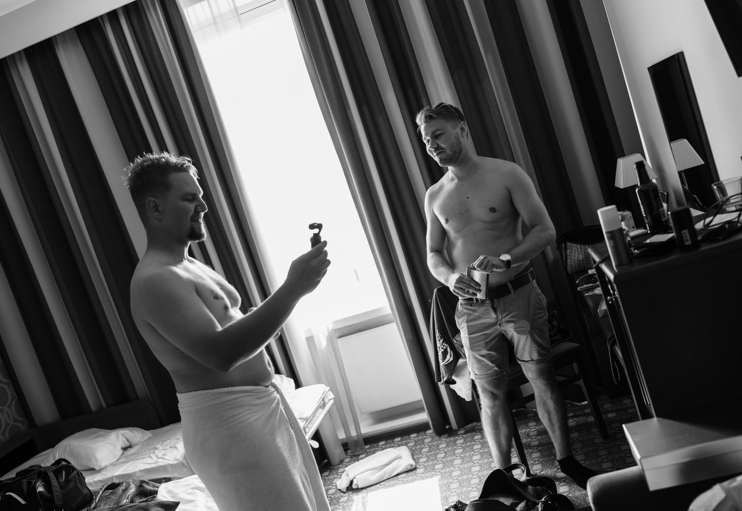 Two shirtless men in a hotel room, one wearing a towel and holding a smartphone, the other wearing shorts and holding an object, with a cluttered background.
