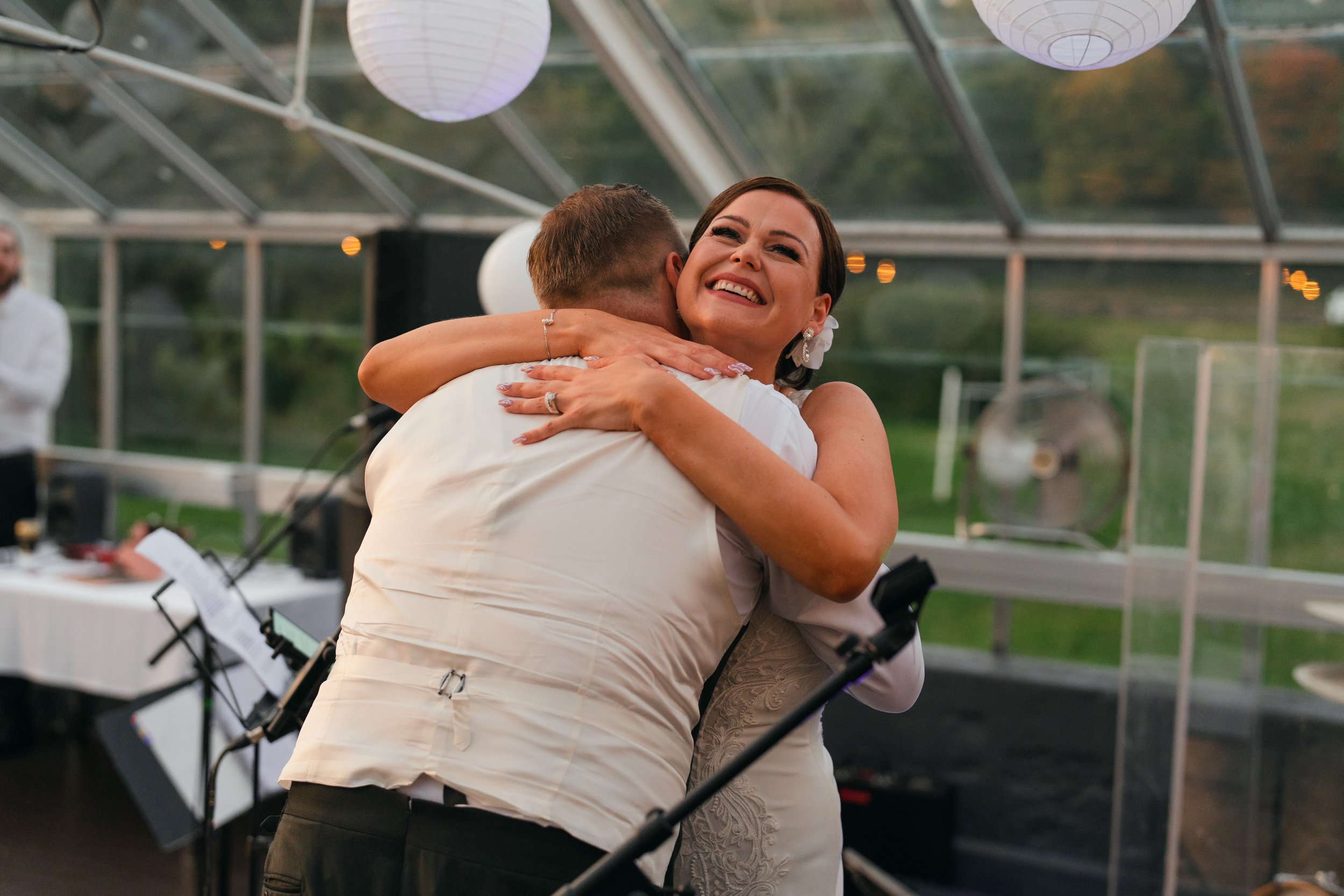 A couple hugging at a wedding reception inside a greenhouse with paper lanterns and a microphone stand nearby.