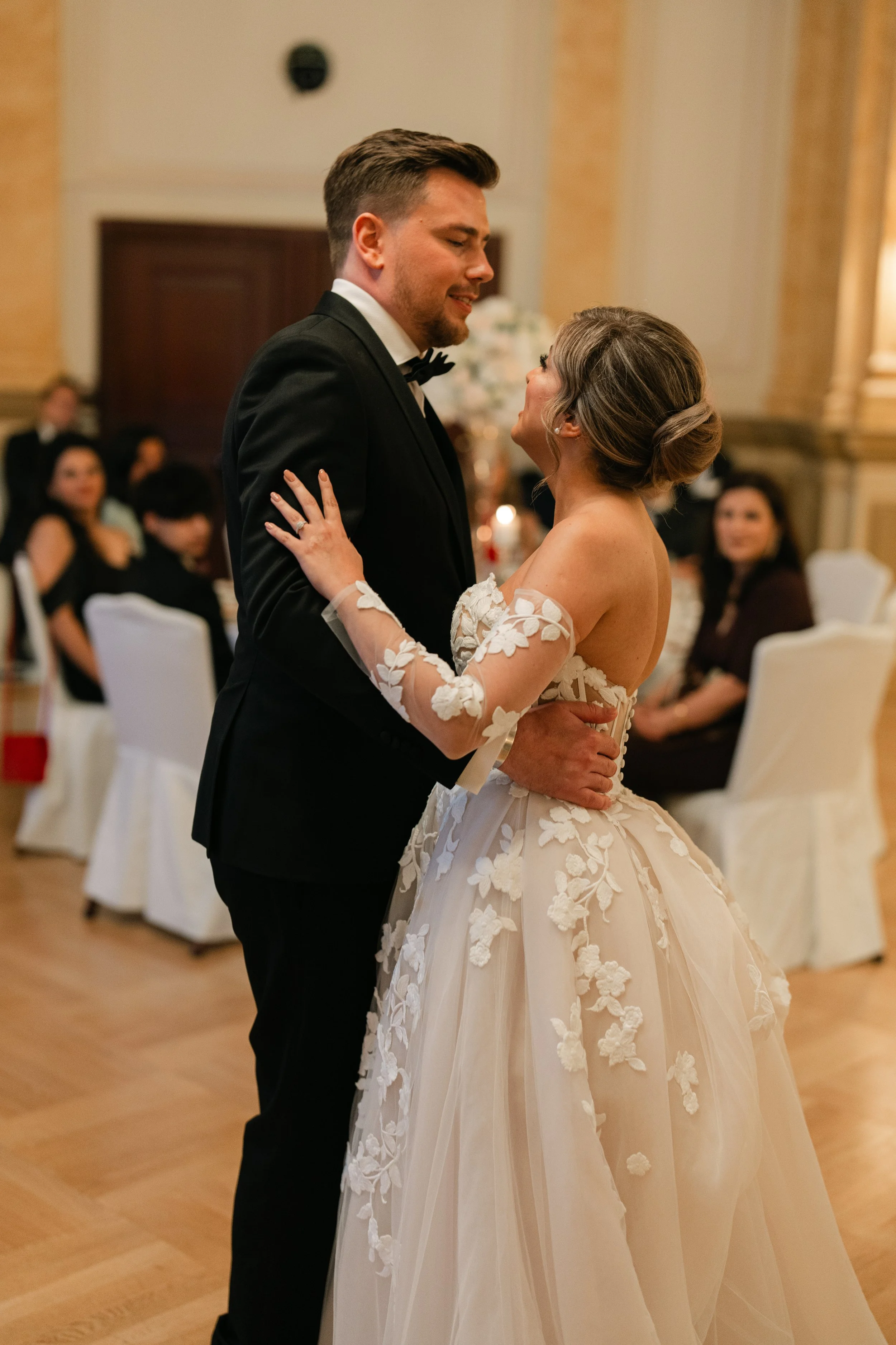 Bride and groom dancing at wedding reception, elegant dress with floral details, guests watching in background.