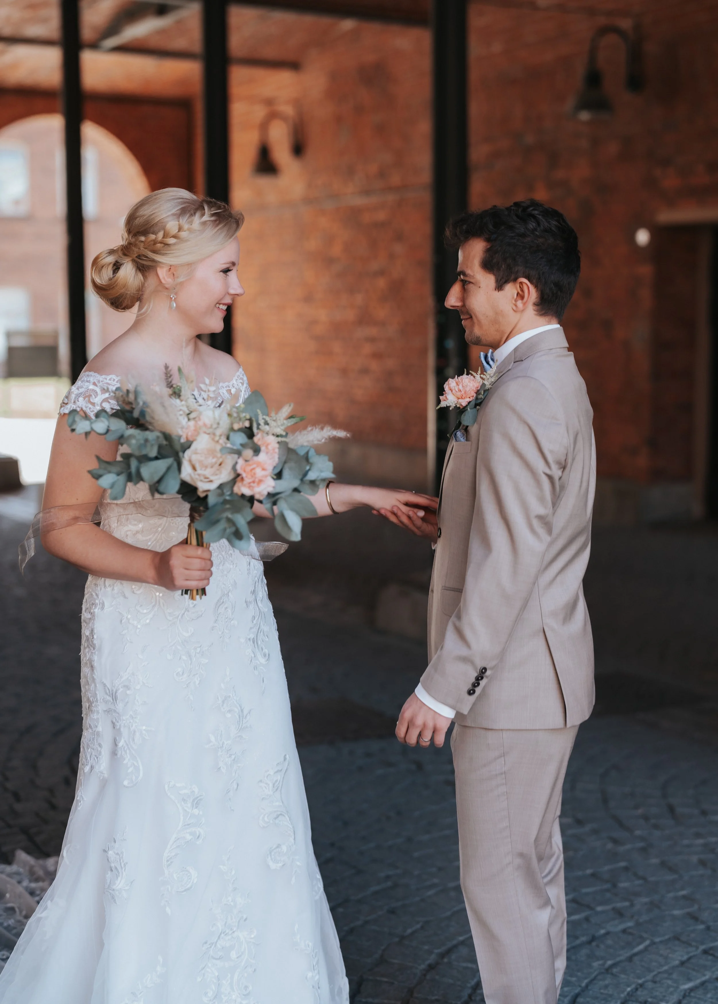 Bride and groom smiling at each other holding hands, with bride holding a bouquet, in an outdoor setting with brick walls.