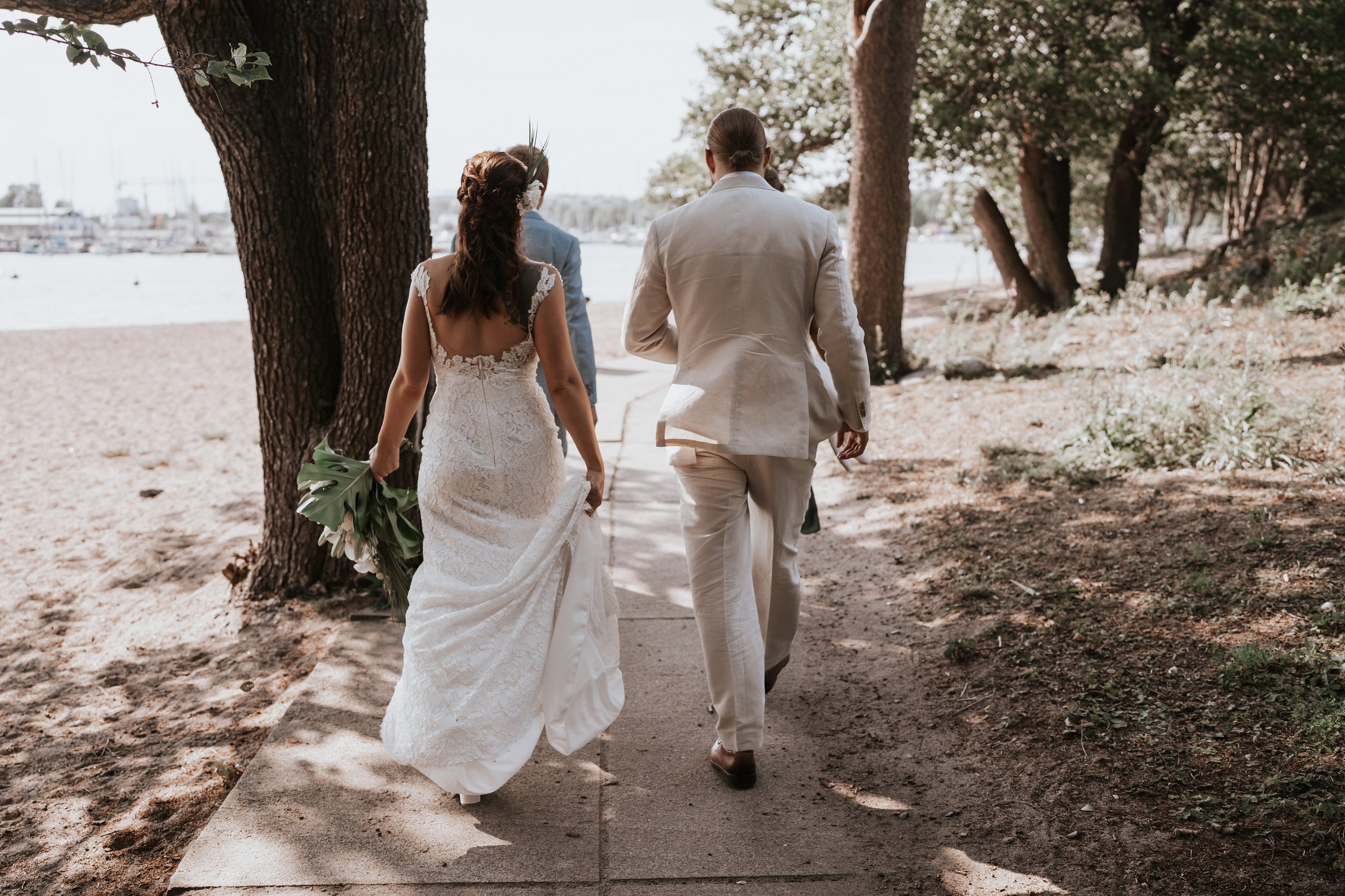 Bride and groom walking on a sandy path near a beach, surrounded by trees.