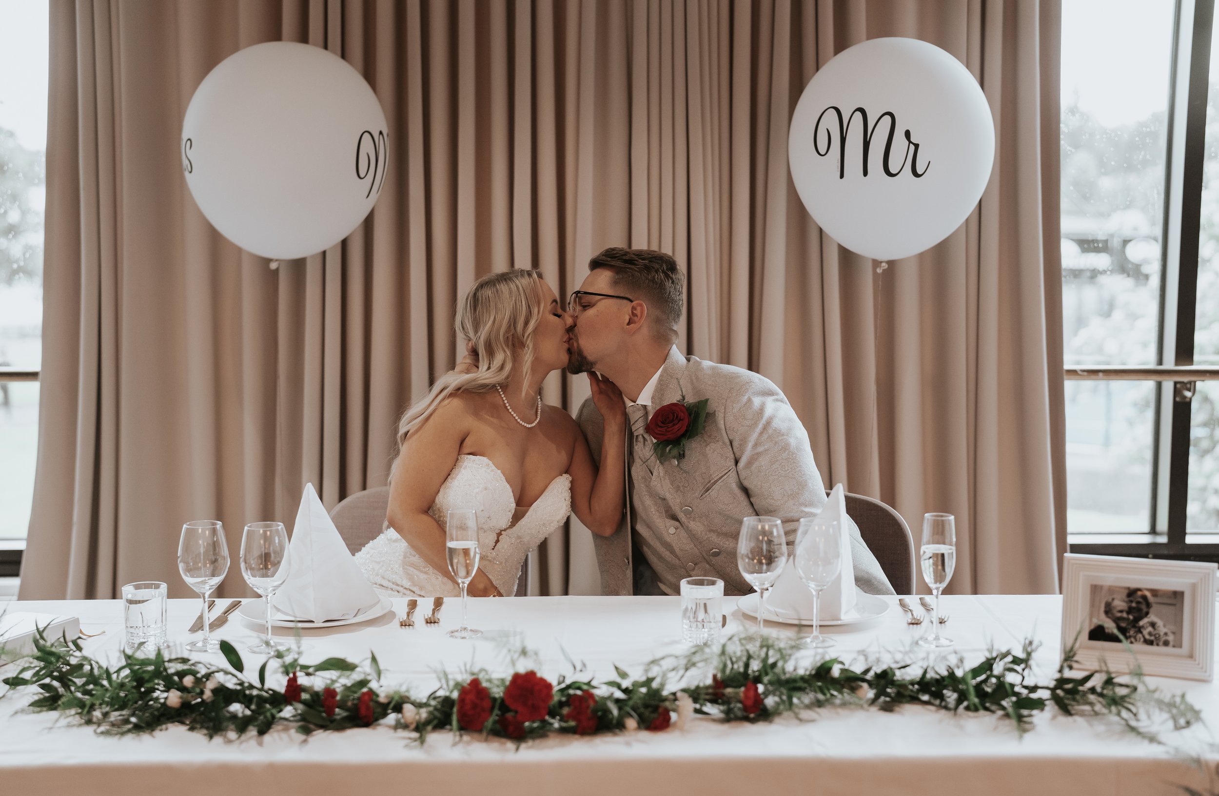 Bride and groom kissing at a wedding reception table, decorated with red roses and greenery, with Mr and Mrs balloons in the background.