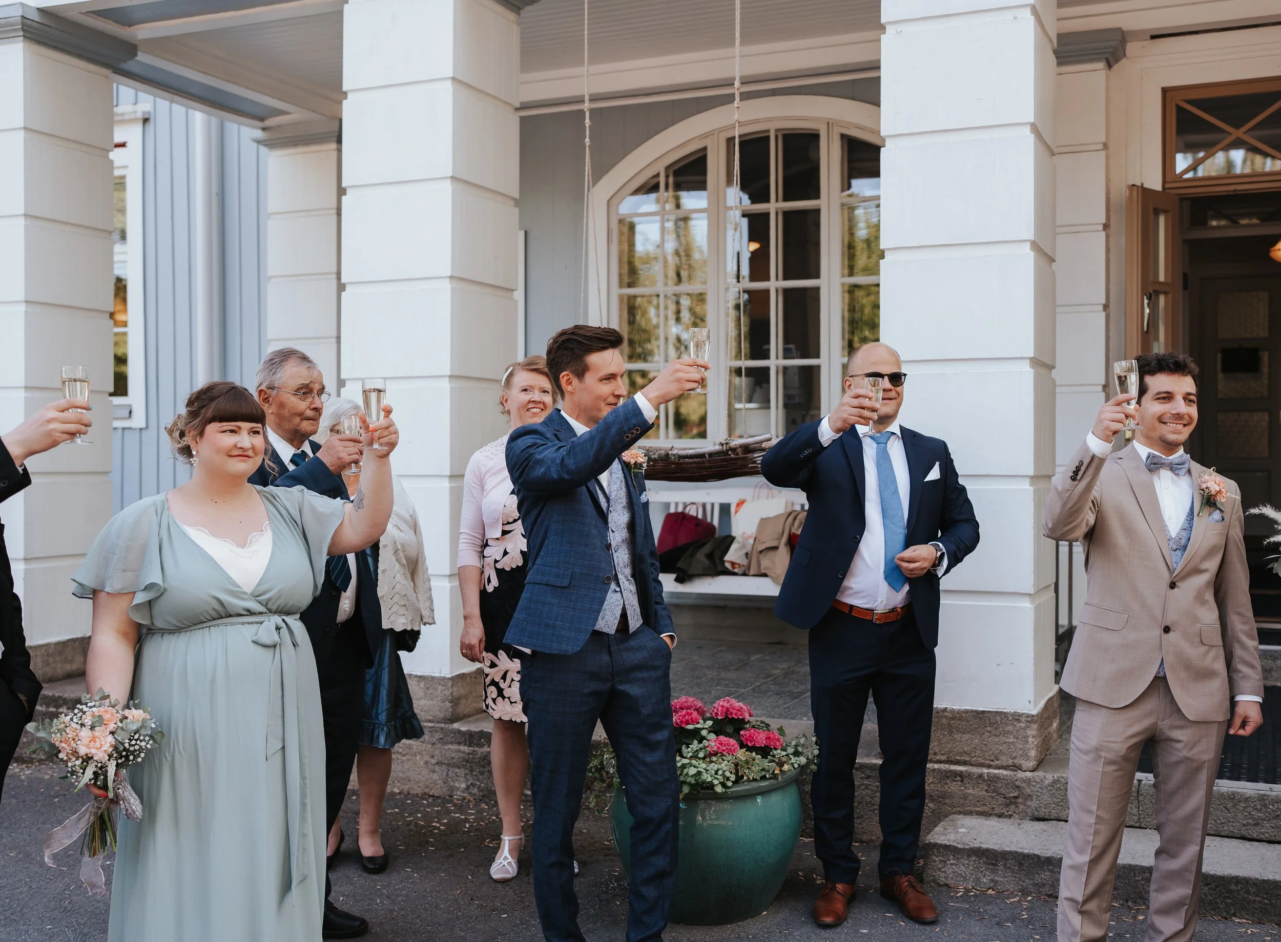 People raising glasses in a toast at an outdoor event, including a woman holding a bouquet, with white columns and a large window in the background.