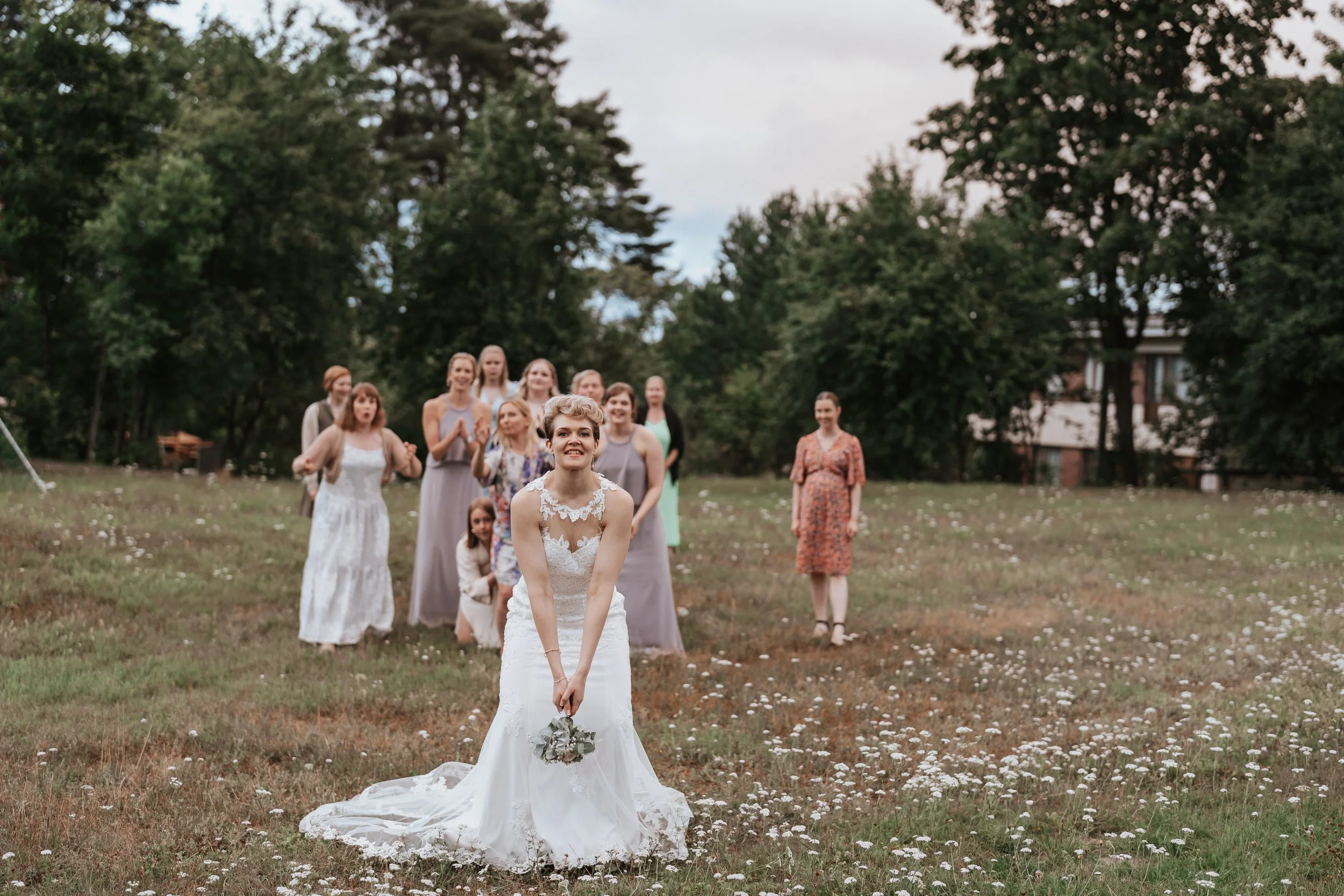Bride in a wedding dress holding bouquet, preparing to toss it to a group of women standing in a grassy field with trees in the background.