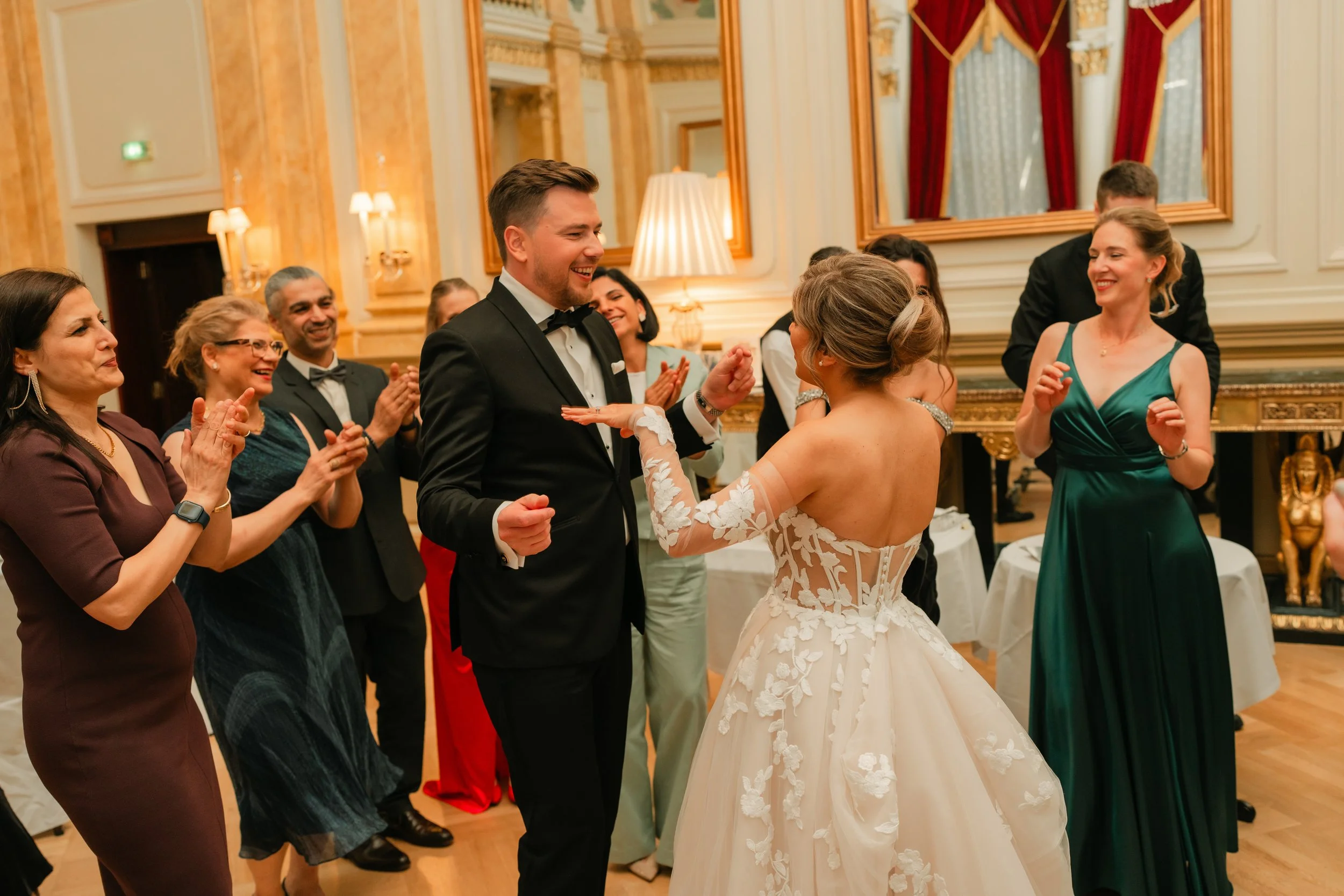 A bride in a white floral gown and a groom in a tuxedo dance surrounded by smiling guests in an elegant ballroom. The guests are clapping and wearing formal attire. The setting features luxurious decor with gold and red accents.