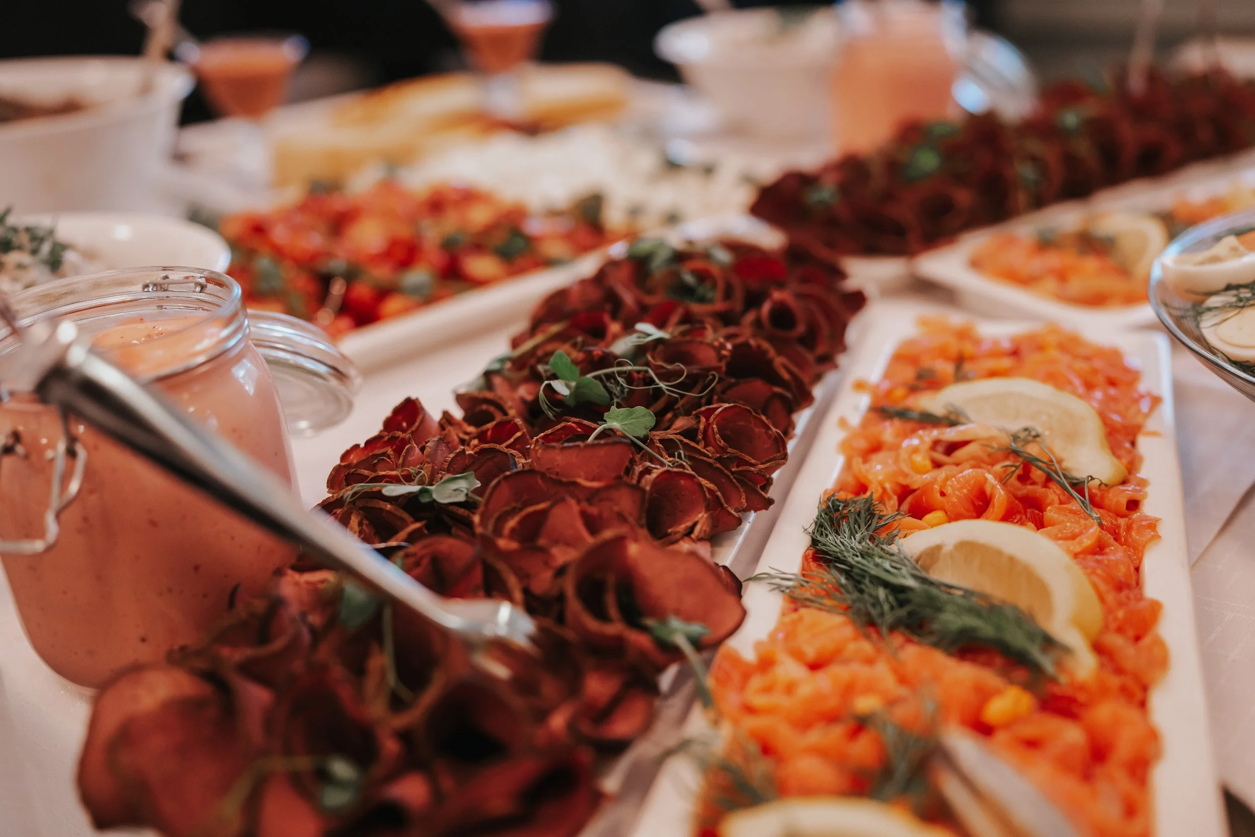 Buffet table with assorted dishes including cured meats, salmon with lemon and herbs, and a jar of sauce.