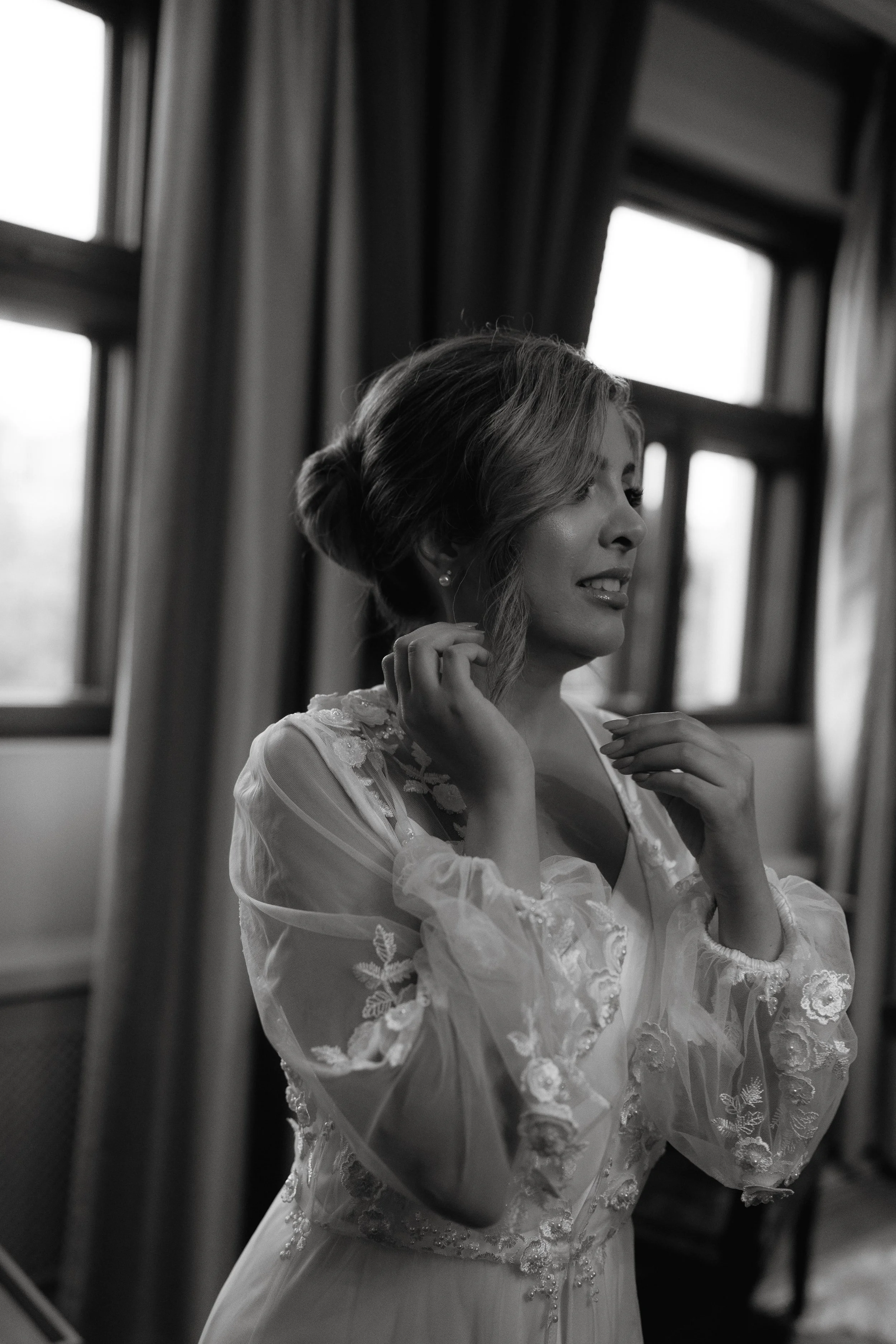 Black and white photo of a bride in a lace robe standing near a window