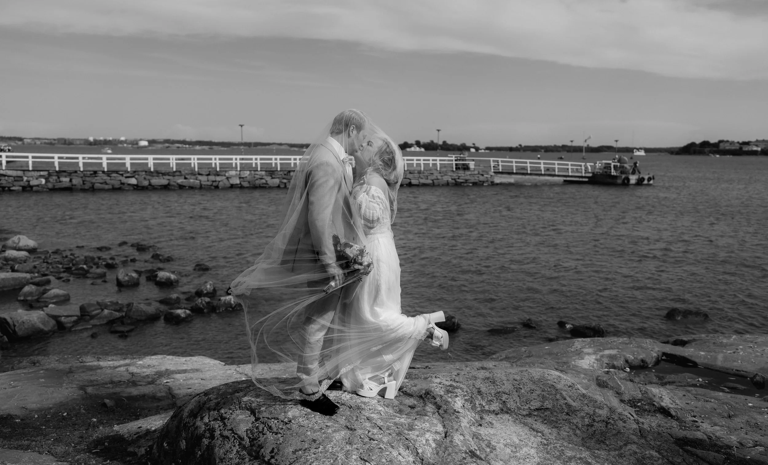 Bride and groom kissing on rocky shore with a veil, waterfront background.