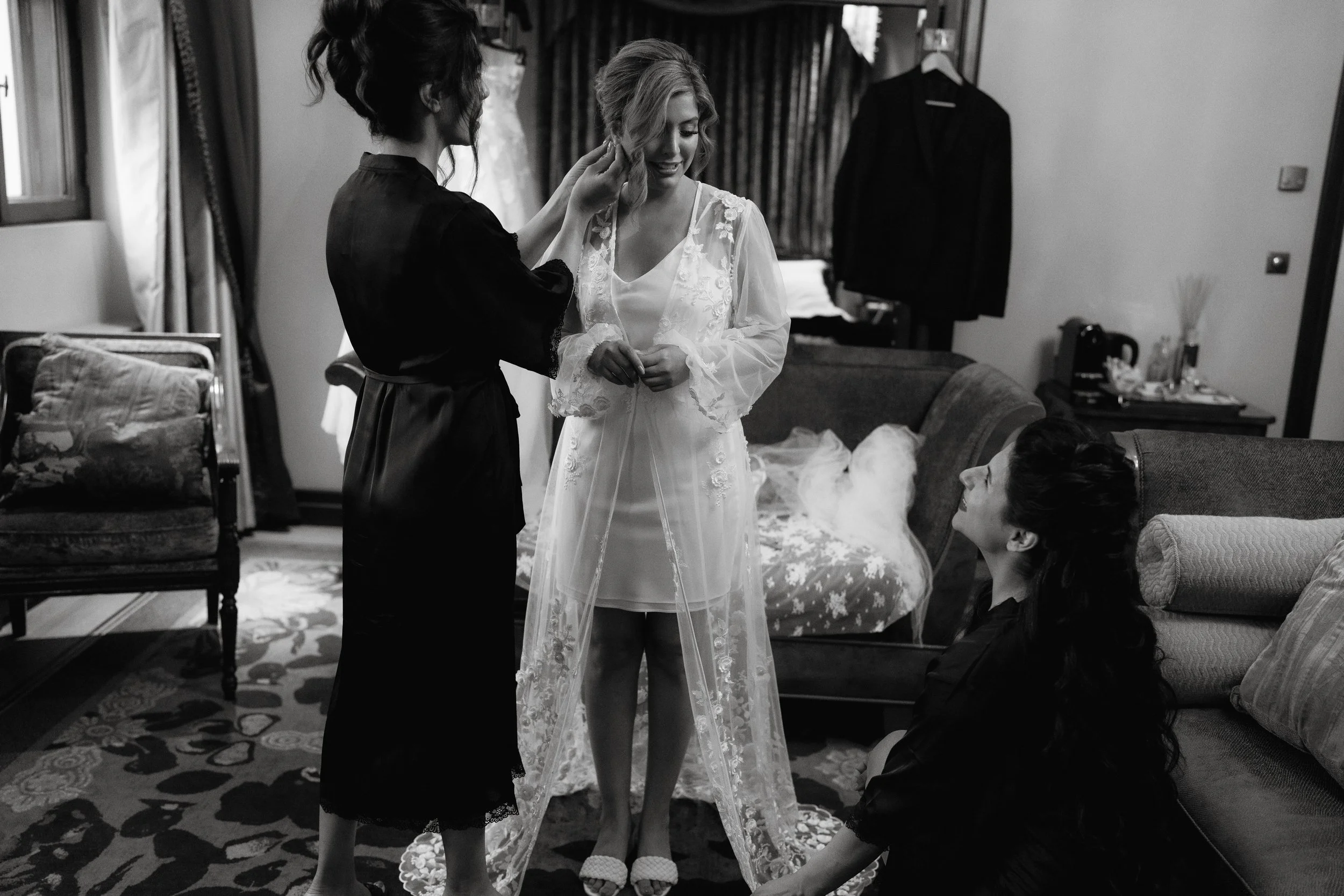 Bride getting ready with bridesmaids in a dressing room, wearing a lace robe.