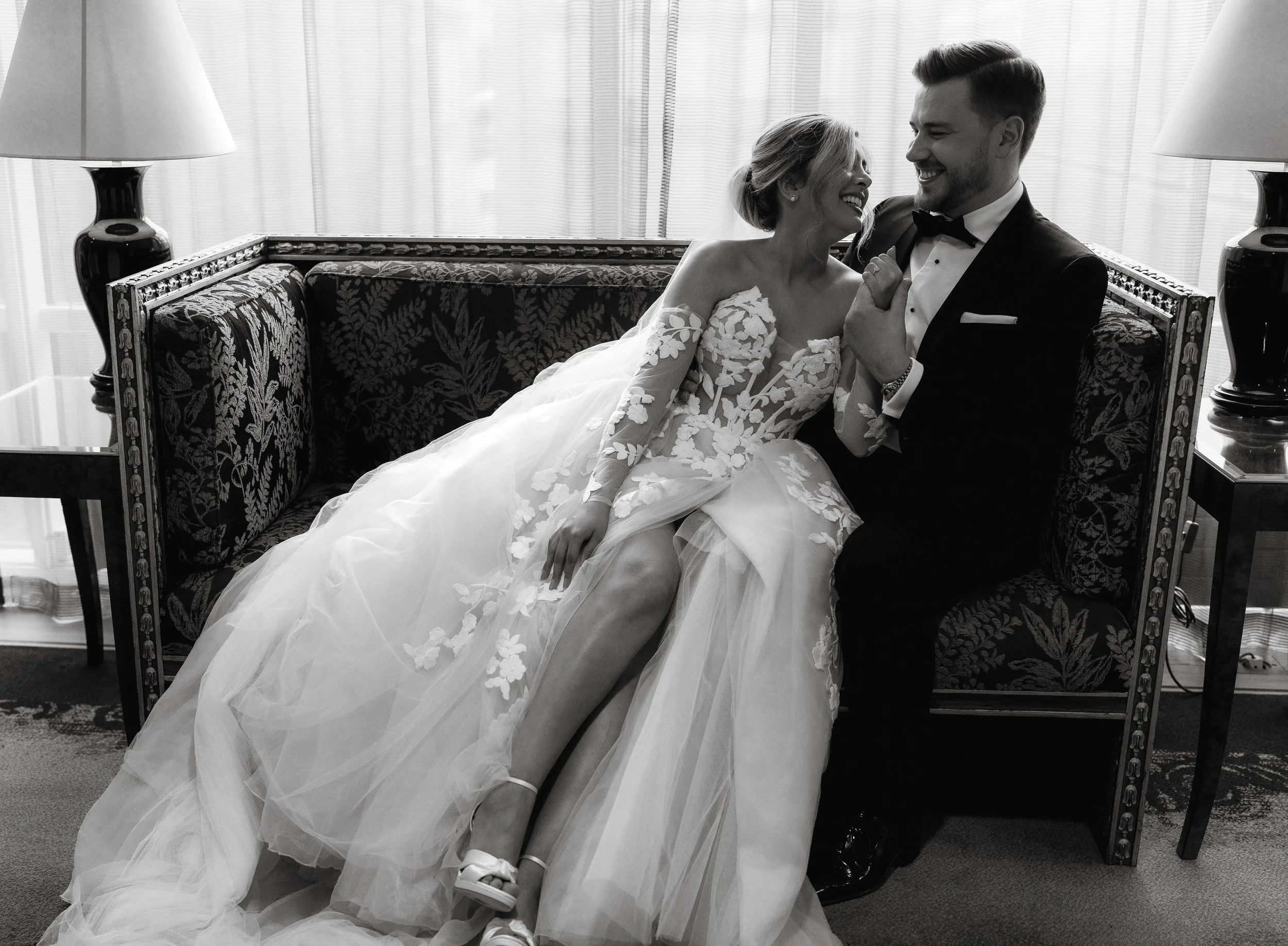 Bride and groom sitting on a sofa, laughing together, in elegant wedding attire; black and white photo.