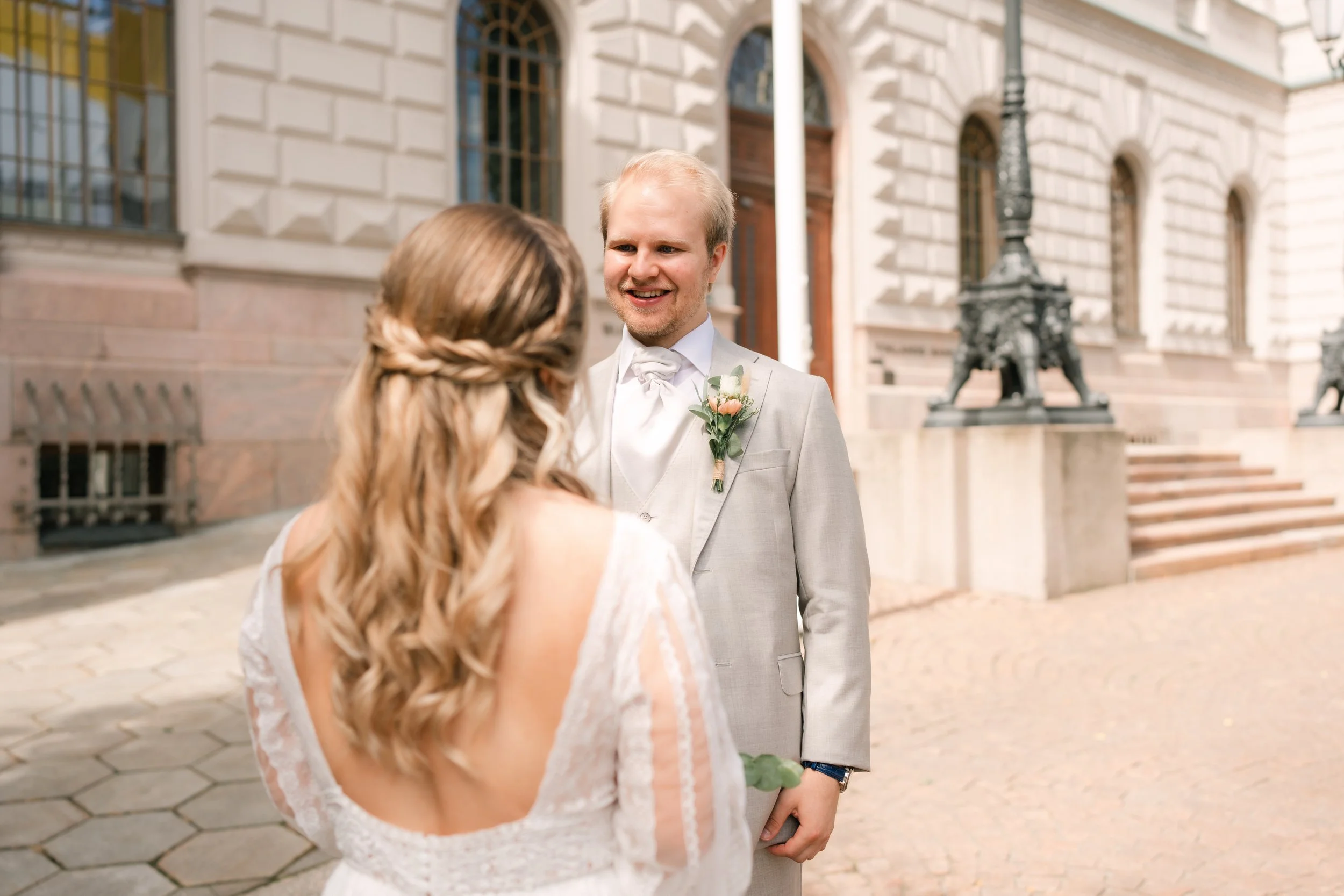 Bride and groom smiling outside a building on their wedding day, with the groom in a light gray suit and the bride in a lace wedding dress, sunny day.